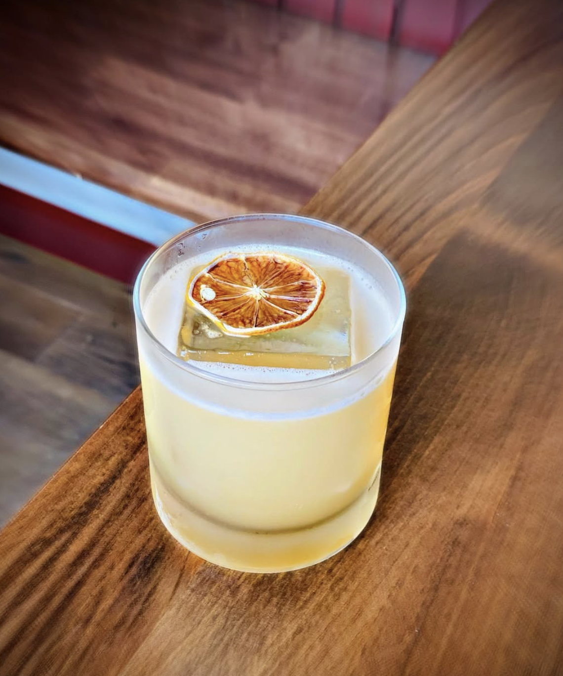 A rocks glass containing a light yellow drink with a large clear cocktail ice cube and a slice of dried orange on top, placed on a wooden surface.