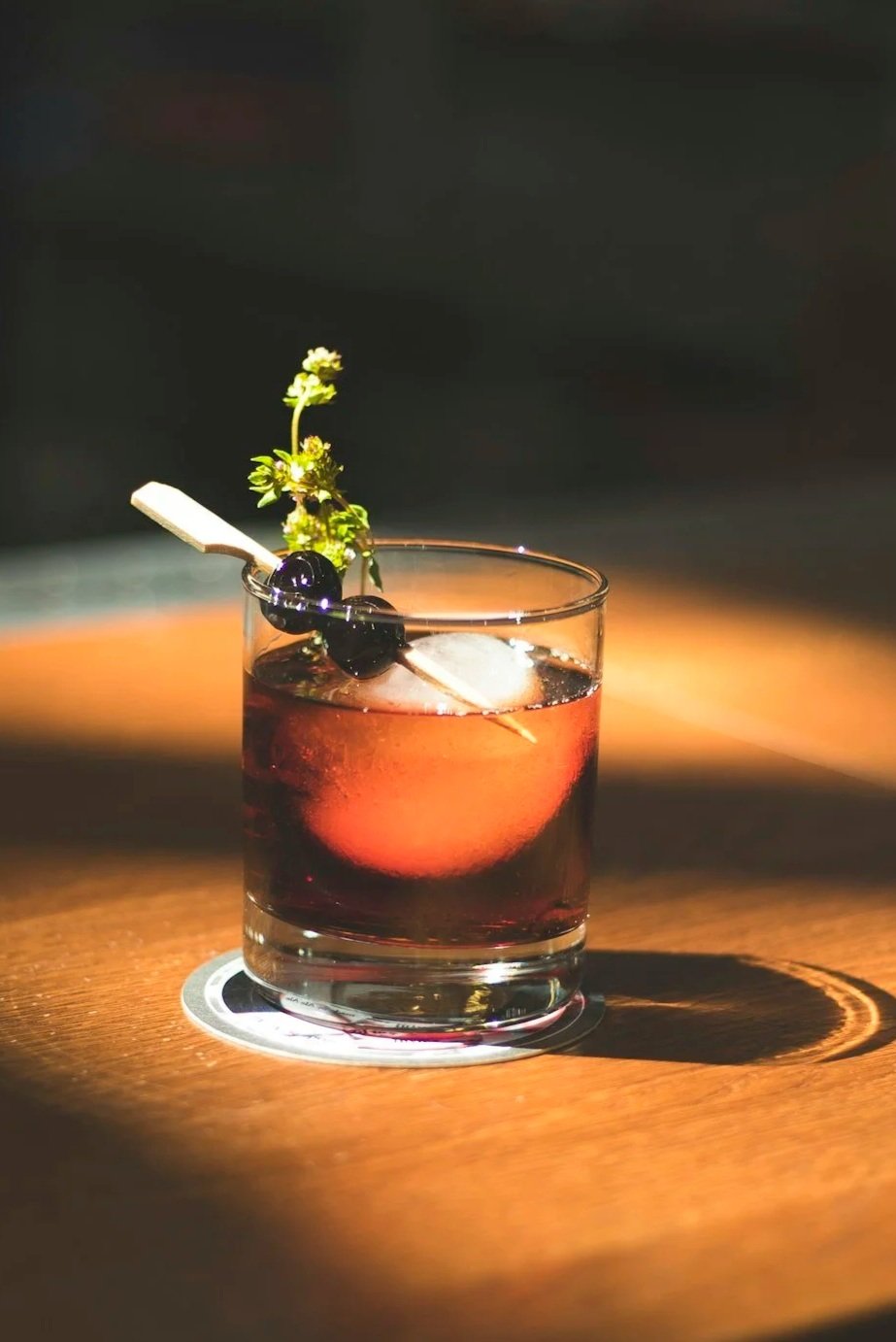 A cocktail in a glass with a big clear ice cube, dark red liquid, garnished with blueberries and a sprig of herbs, on a wooden surface.