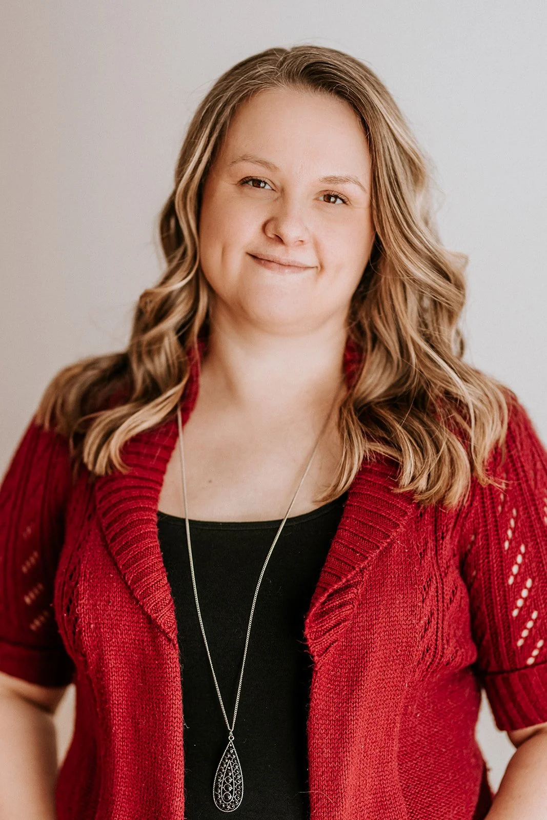 A woman with dark blond, wavy hair wearing a red knit cardigan, black top, and a silver necklace with a teardrop pendant, smiling softly against a neutral background.