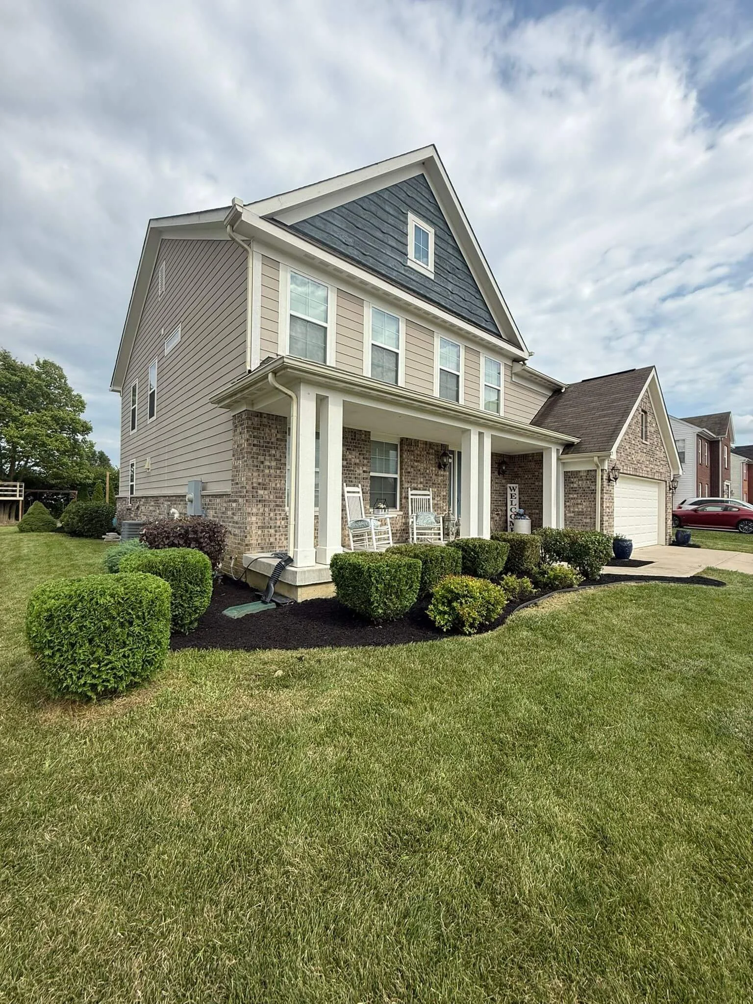 Front view of a two-story suburban house with a brick and beige siding exterior, white porch, green lawn, and neatly trimmed bushes. The sky is partly cloudy.