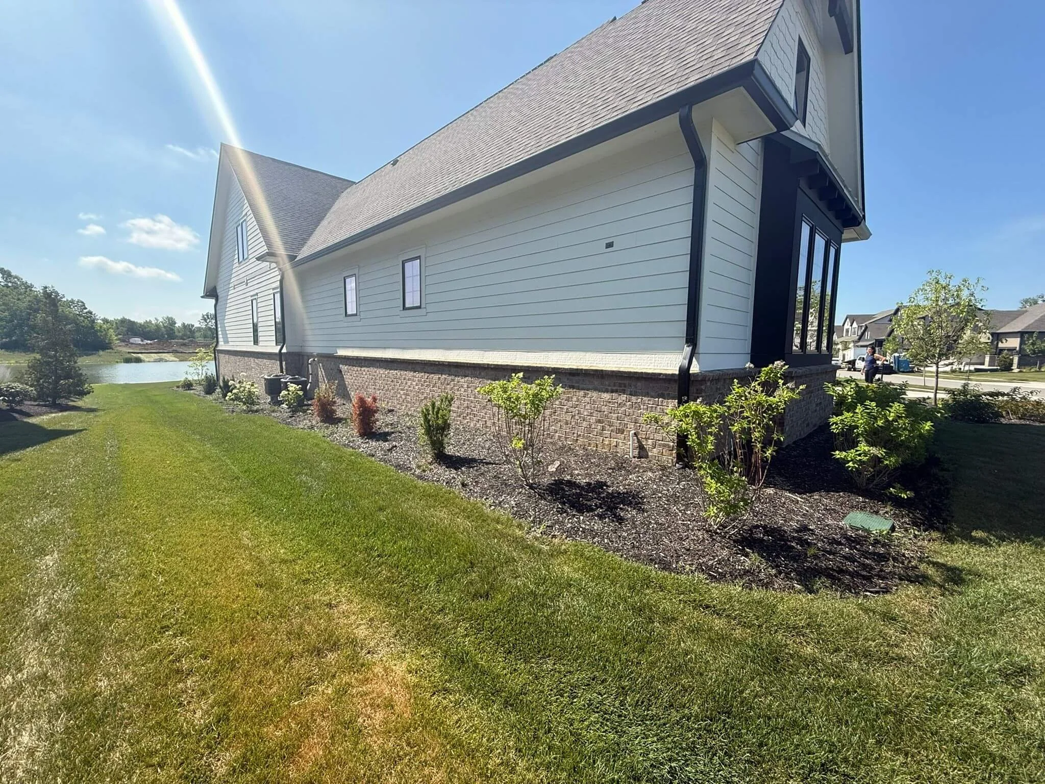 Side view of a modern house with white siding, black trim, and a brick foundation, surrounded by a well-maintained grassy yard with small shrubs and trees, near a pond on a sunny day.