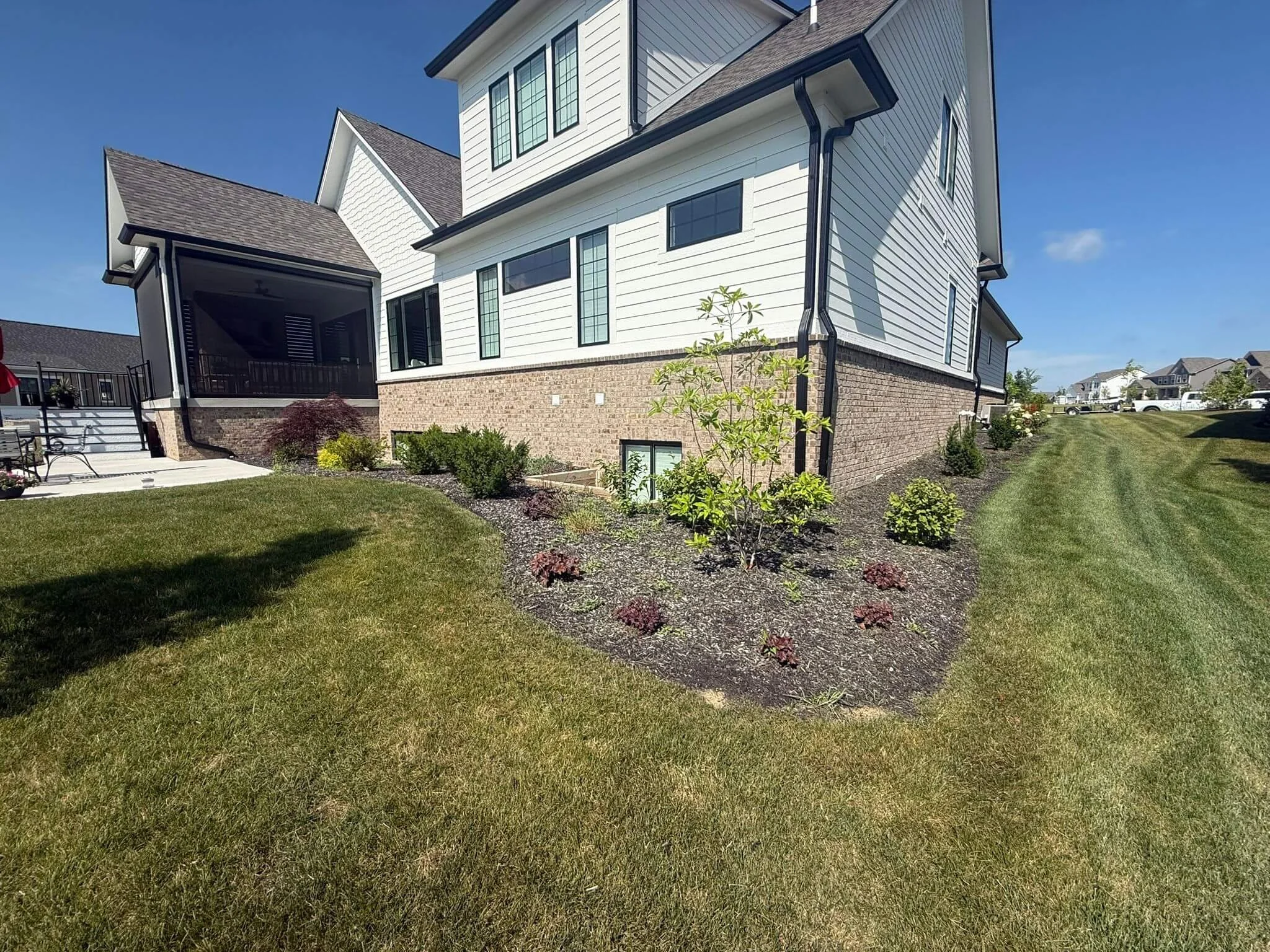 A modern two-story house with white siding and brick foundation, surrounded by a well-maintained lawn and landscaped garden with small shrubs and young trees. There is a screened porch on the left side, and the sky is clear with a few clouds.