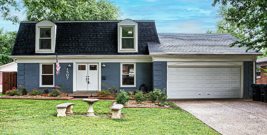 A blue two-story house with dormer windows, a white garage door, and a front yard with a birdbath and benches surrounded by flower beds.