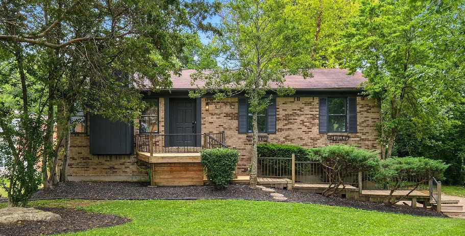 A brick house with a dark door and shutters, surrounded by a landscaped yard with stairs and a small deck, shaded by trees.