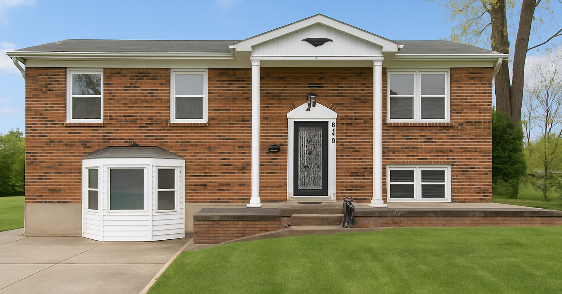 A brick two-story house with white trim, a front porch with columns, a black front door, multiple windows, and a bay window on the left side. There is a well-maintained lawn, trees in the background, and a concrete walkway leading to the steps of the