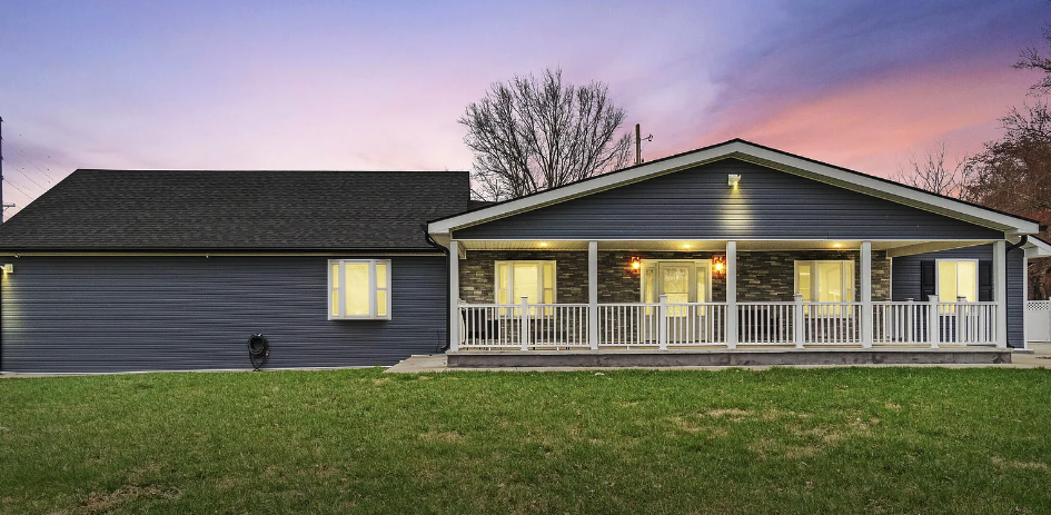 A single-story house with blue siding, a large front porch with white railings, and multiple windows, during twilight with a pinkish sky.