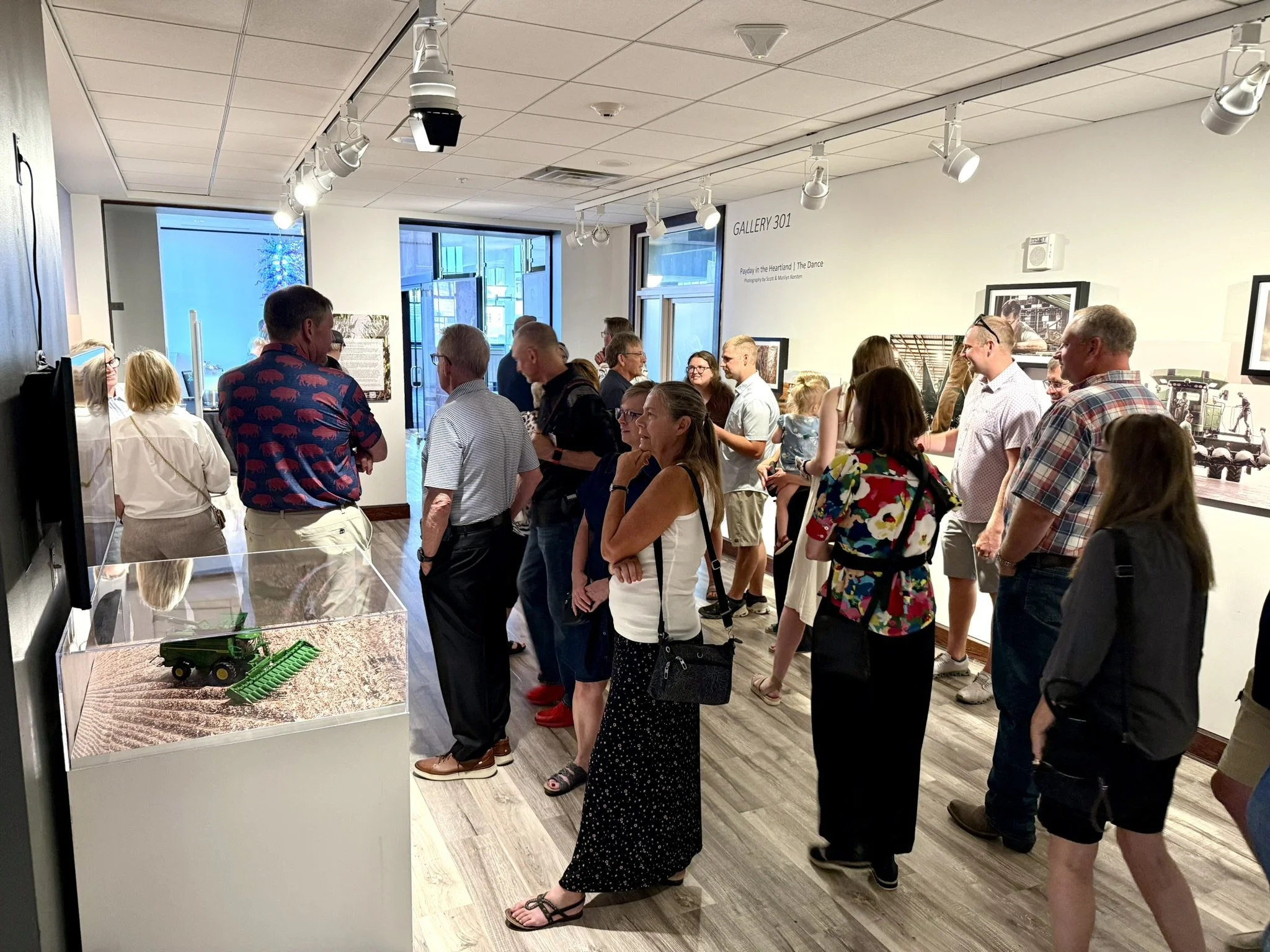 People gathered in an art gallery, viewing framed art photographs and on the walls. The room has a ceiling with multiple light fixtures, and large glass doors let in natural light.