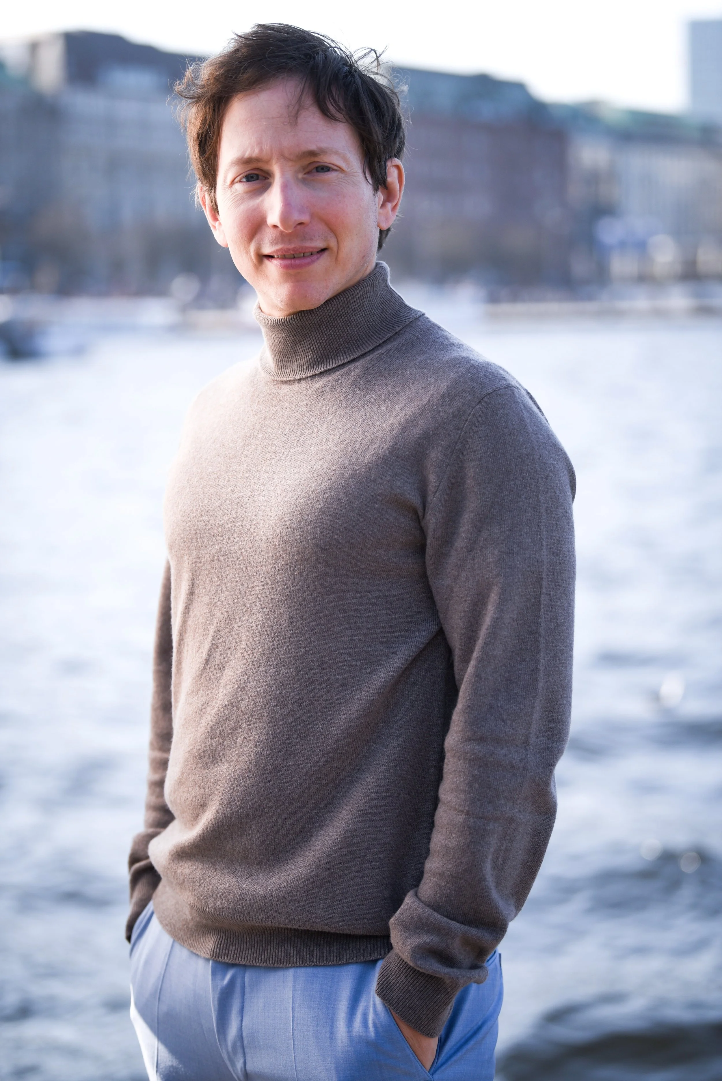 Scharfer Blick eines Mannes in einem braunen Rollkragenpullover, der am Wasser steht, mit einer Stadt im Hintergrund.