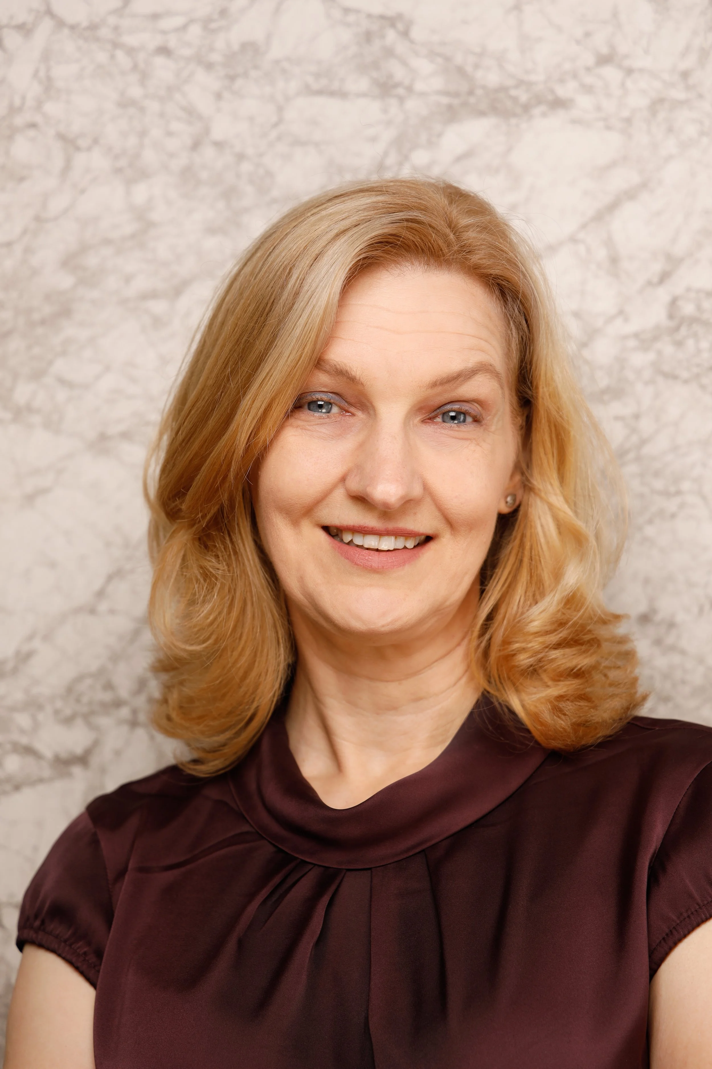 Porträt einer lächelnden Frau mit blonden, lockigen Haaren vor einer hellen Marmorwand.