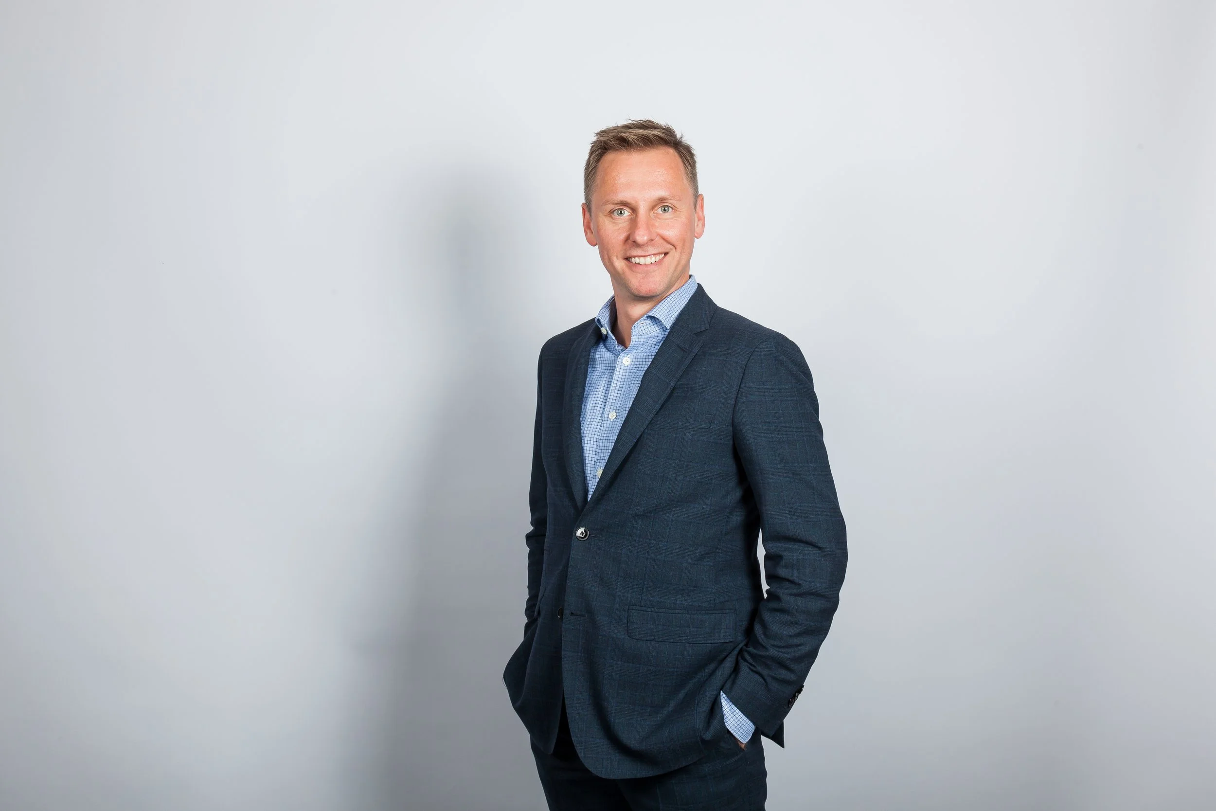Ein Lächelnder Mann in einem dunklen Anzug und blauer Krawatte steht vor einer weißen Wand.