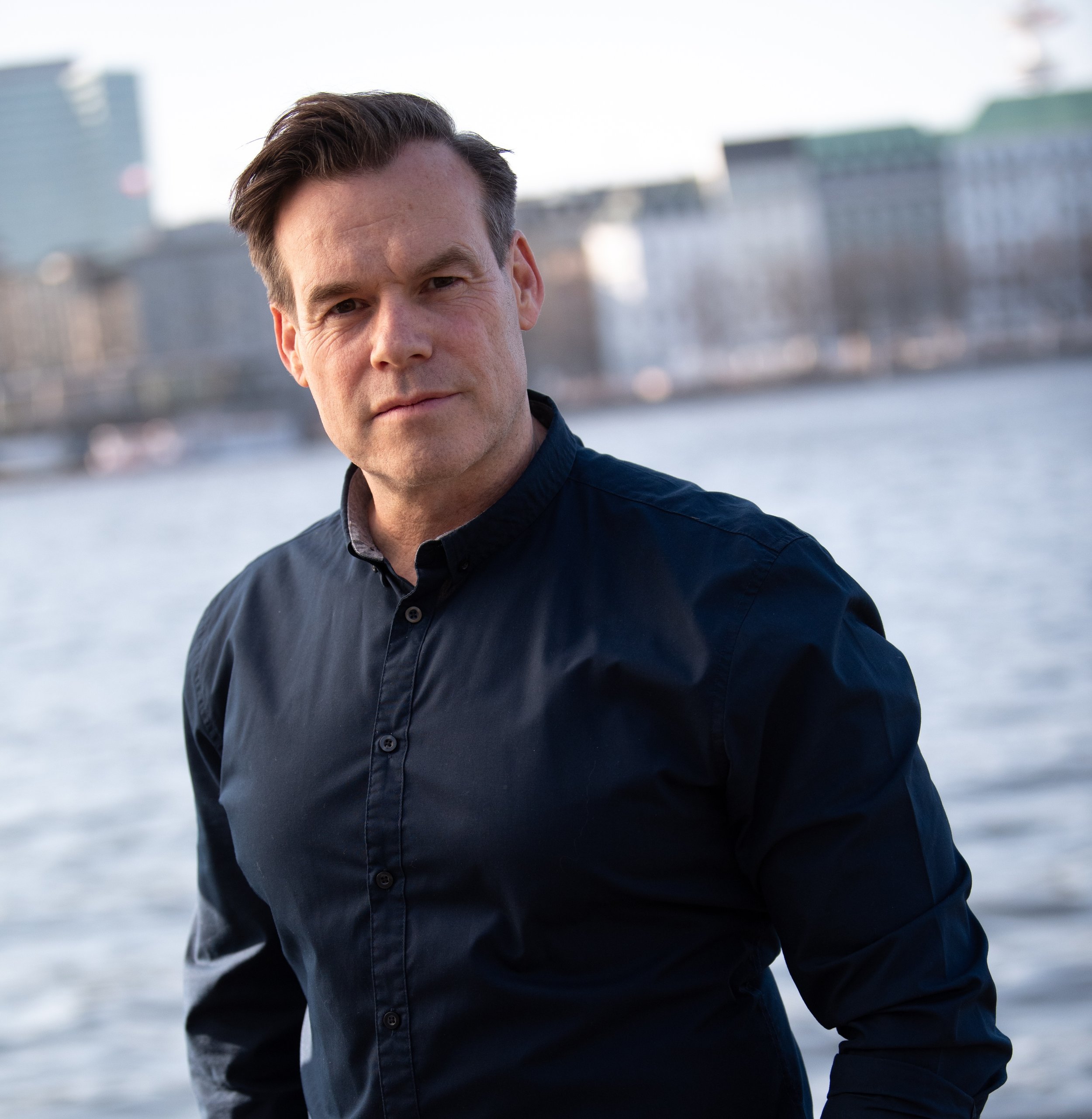 Ein Mann steht in der Nähe eines Flusses, im Hintergrund sind Gebäude zu sehen.