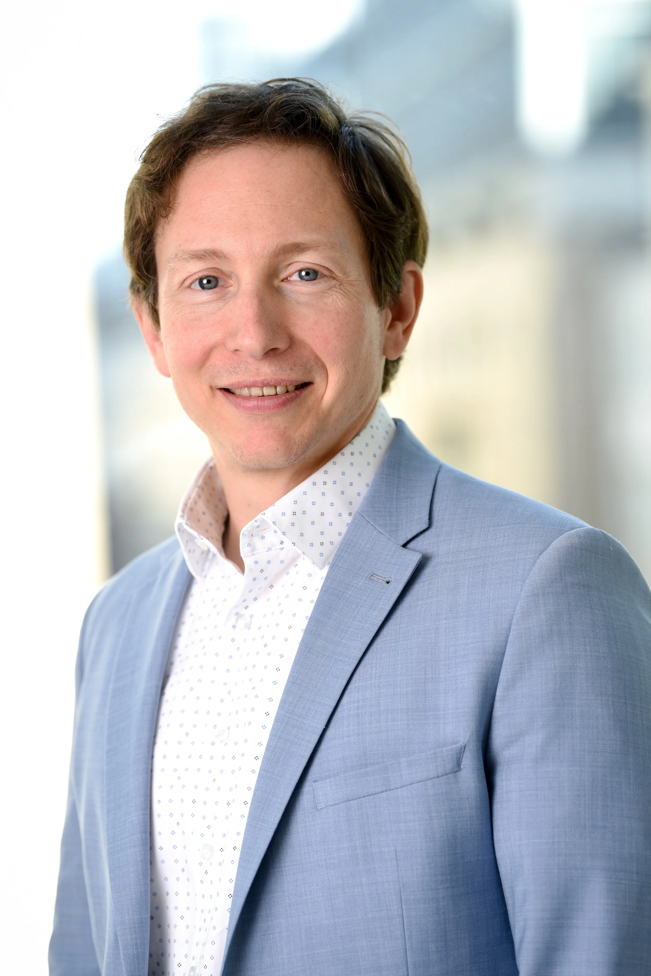 Porträt eines lächelnden Mannes mit braunen Haaren in einem hellblauen Anzug und weißem Hemd, stehend in einem modernen Bürogebäude.