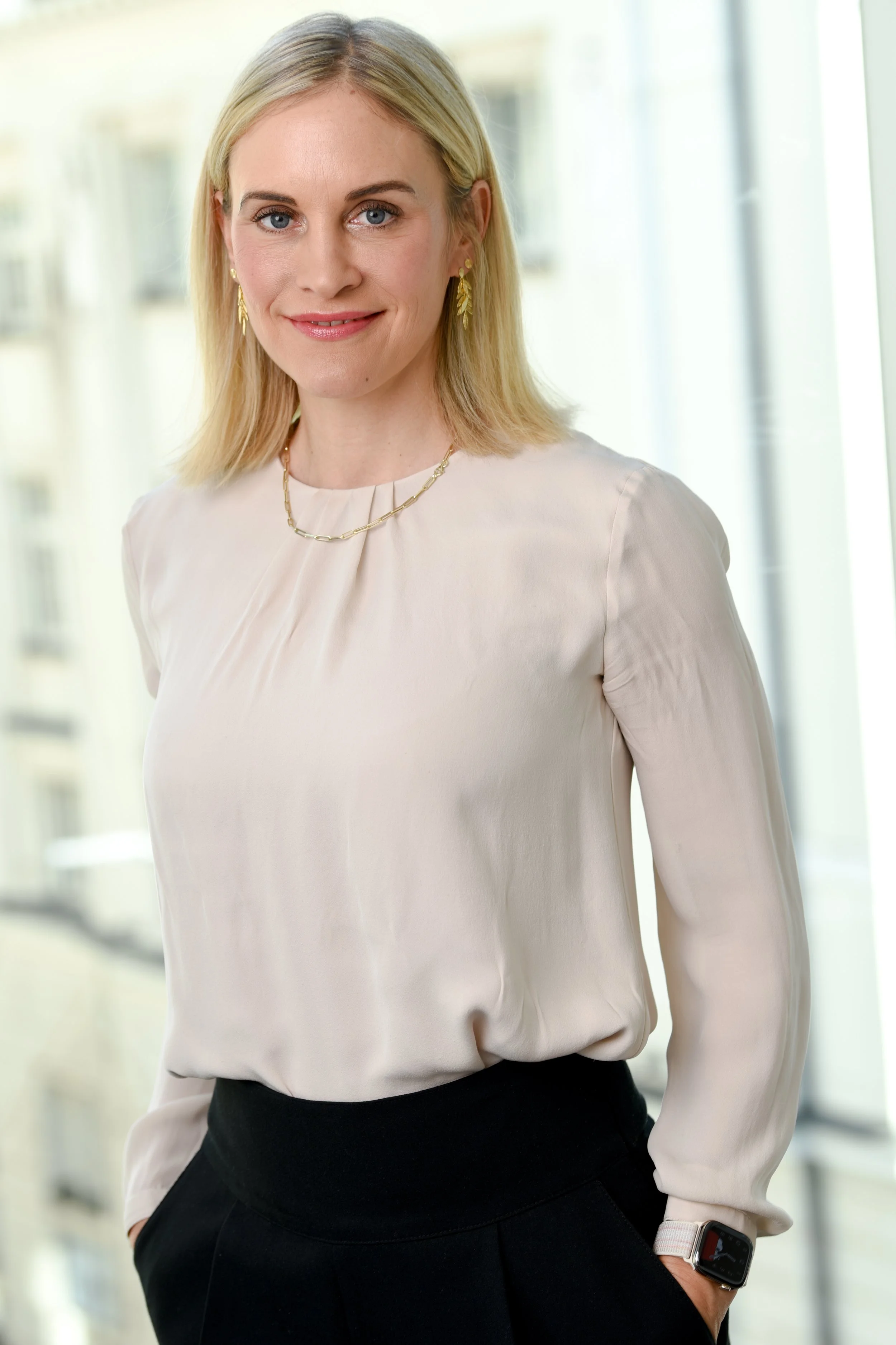 Eine blonde Frau in einem weißen Bluse und schwarzen Hosen steht vor einem großen Fenster in einem modernen Gebäude.