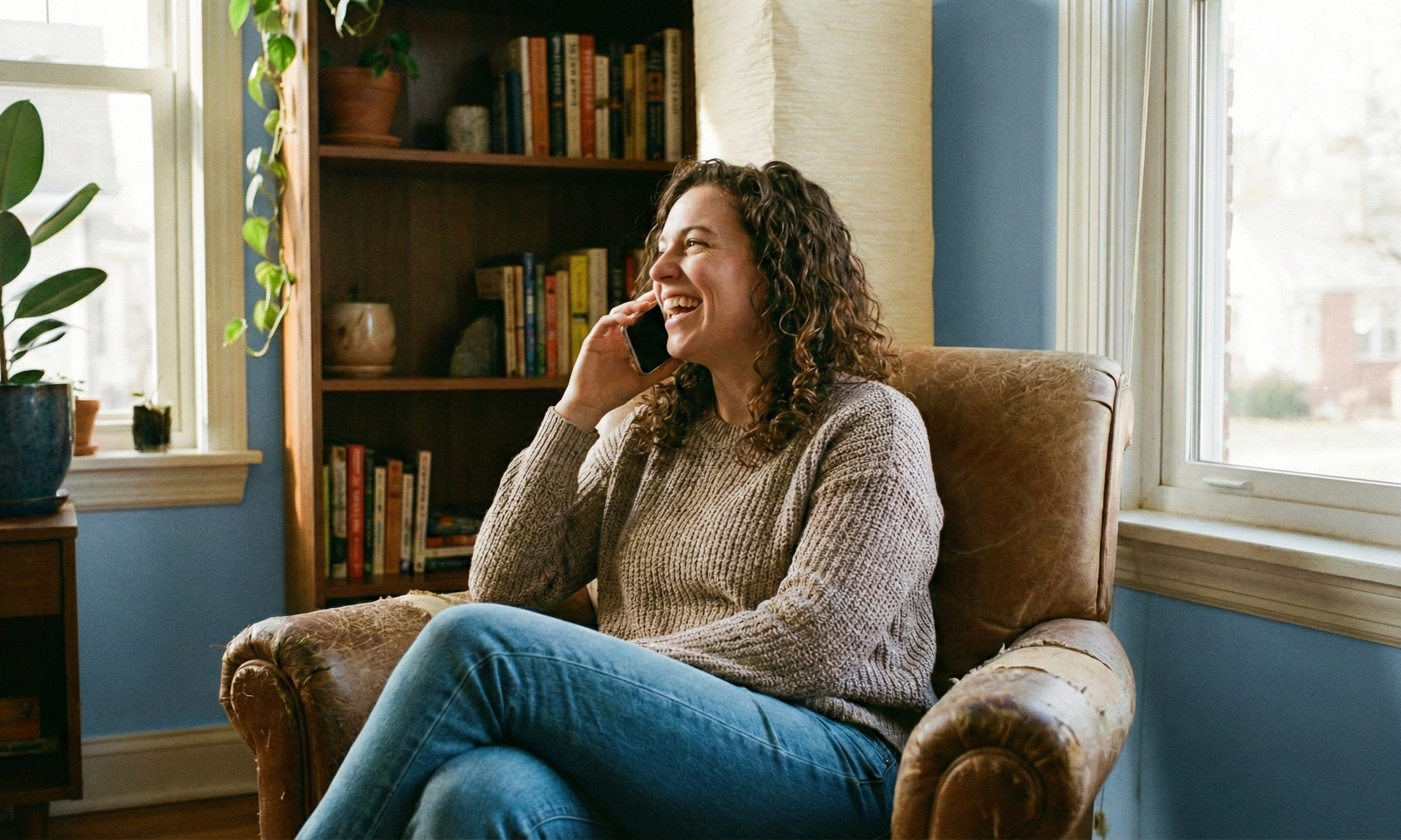 A woman with curly hair smiling and talking on her cellphone while sitting in a comfortable armchair in a cozy living room, with a bookshelf and window in the background.