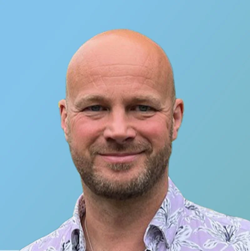 Portrait of a smiling bald man with a beard, wearing a patterned shirt, against a blue background.