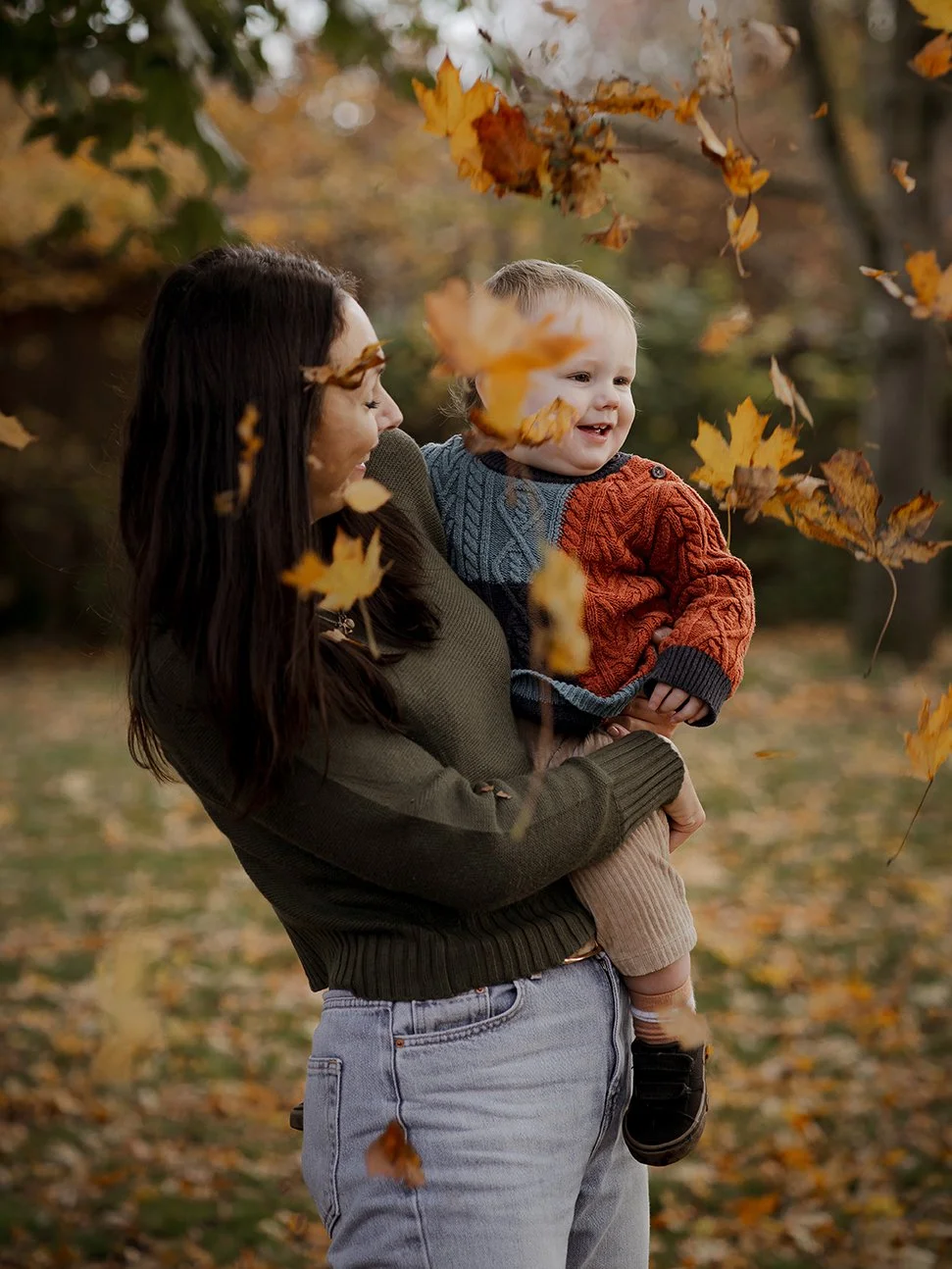 Mother holding her one-year-old son as he laughs in the autumn leaves during a family photoshoot in Henleaze.