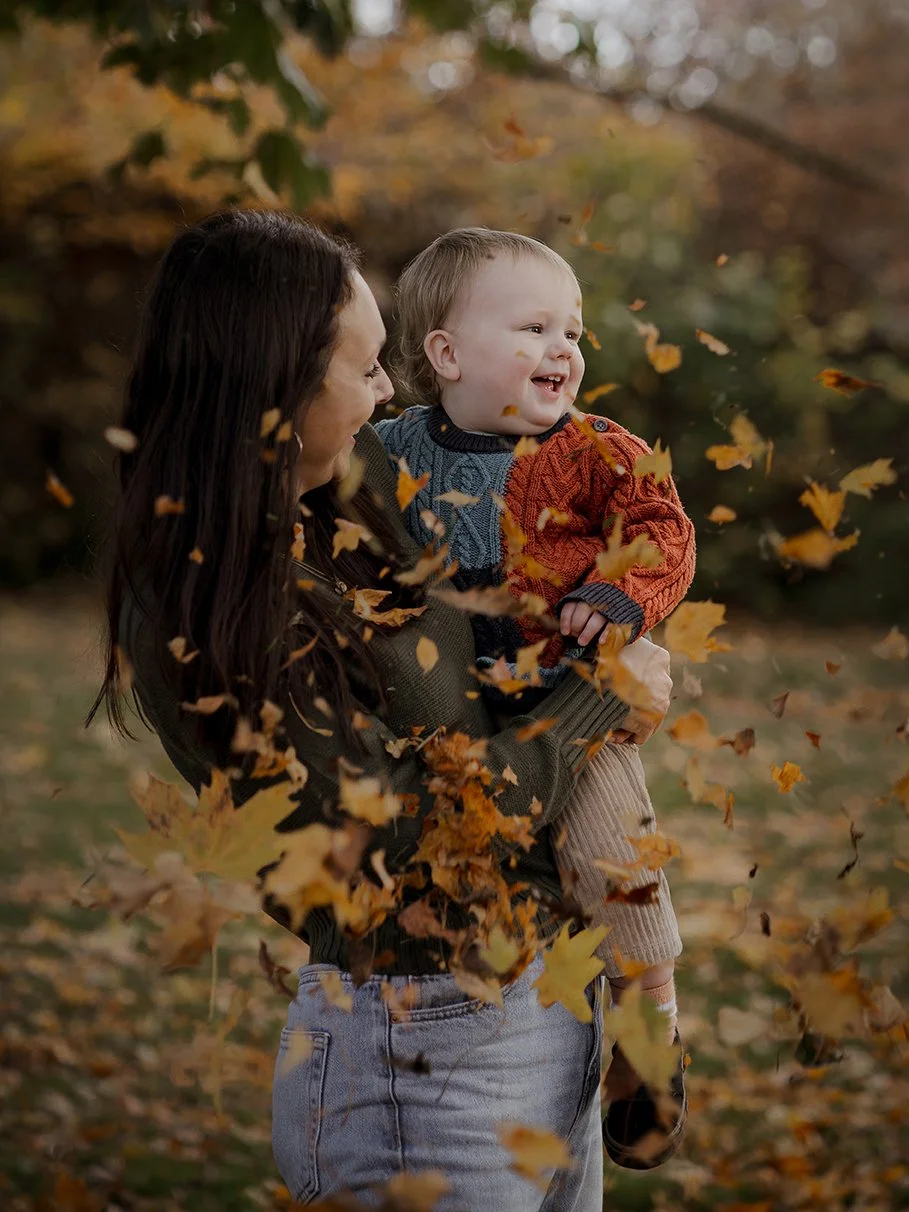 Mother holding her one-year-old son as he laughs in the autumn leaves during a family photoshoot in Henleaze.