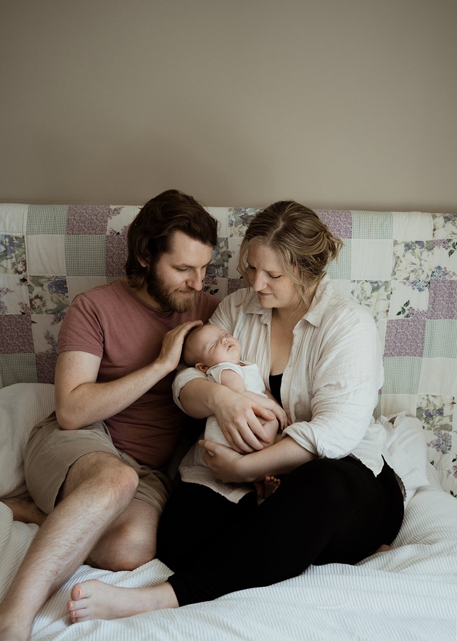 Newborn photographer Bristol - image of a couple with their newborn baby sitting on their bed during a baby photoshoot at their home in Bristol.