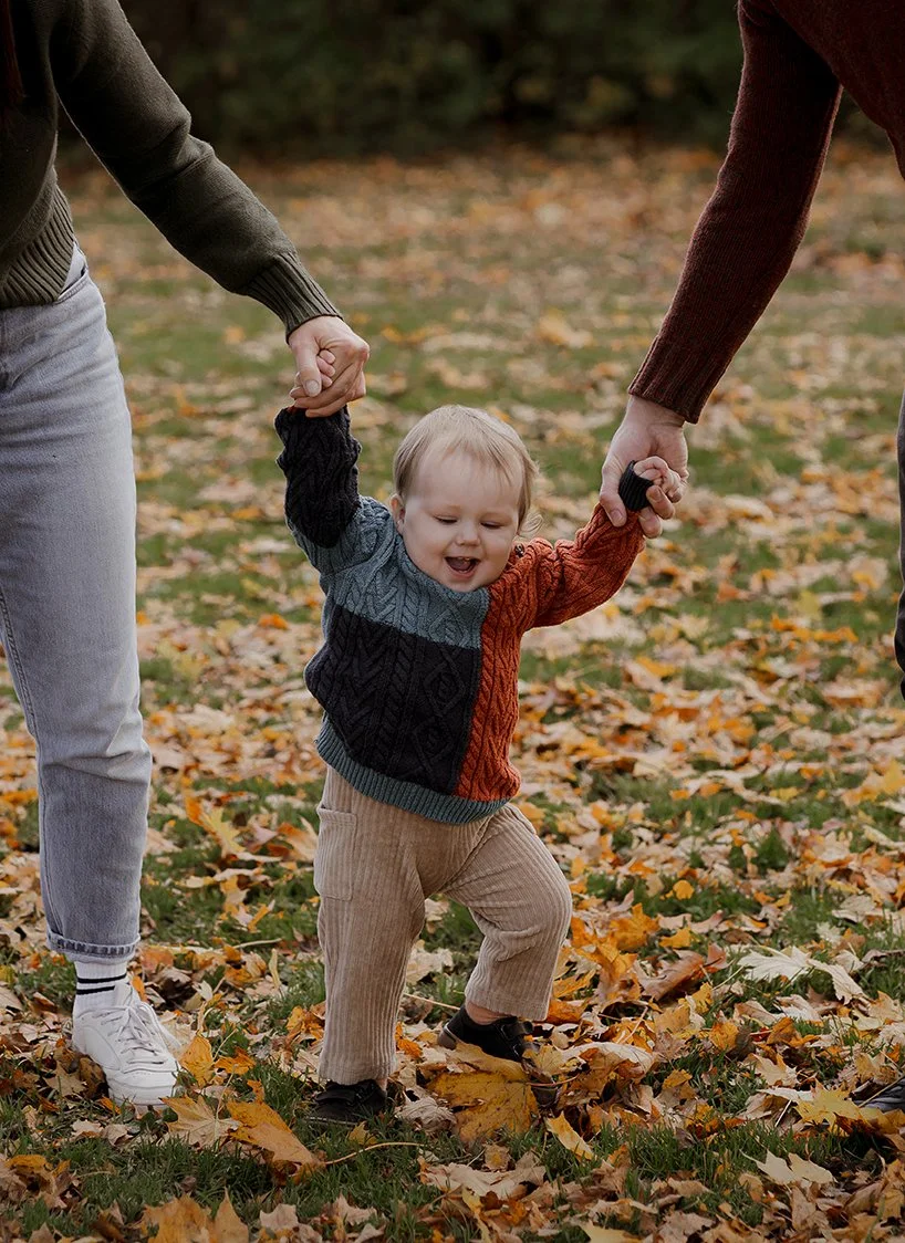 One-year-old boy practising his first steps while holding his parents’ hands during a family photoshoot in Henleaze, Bristol.