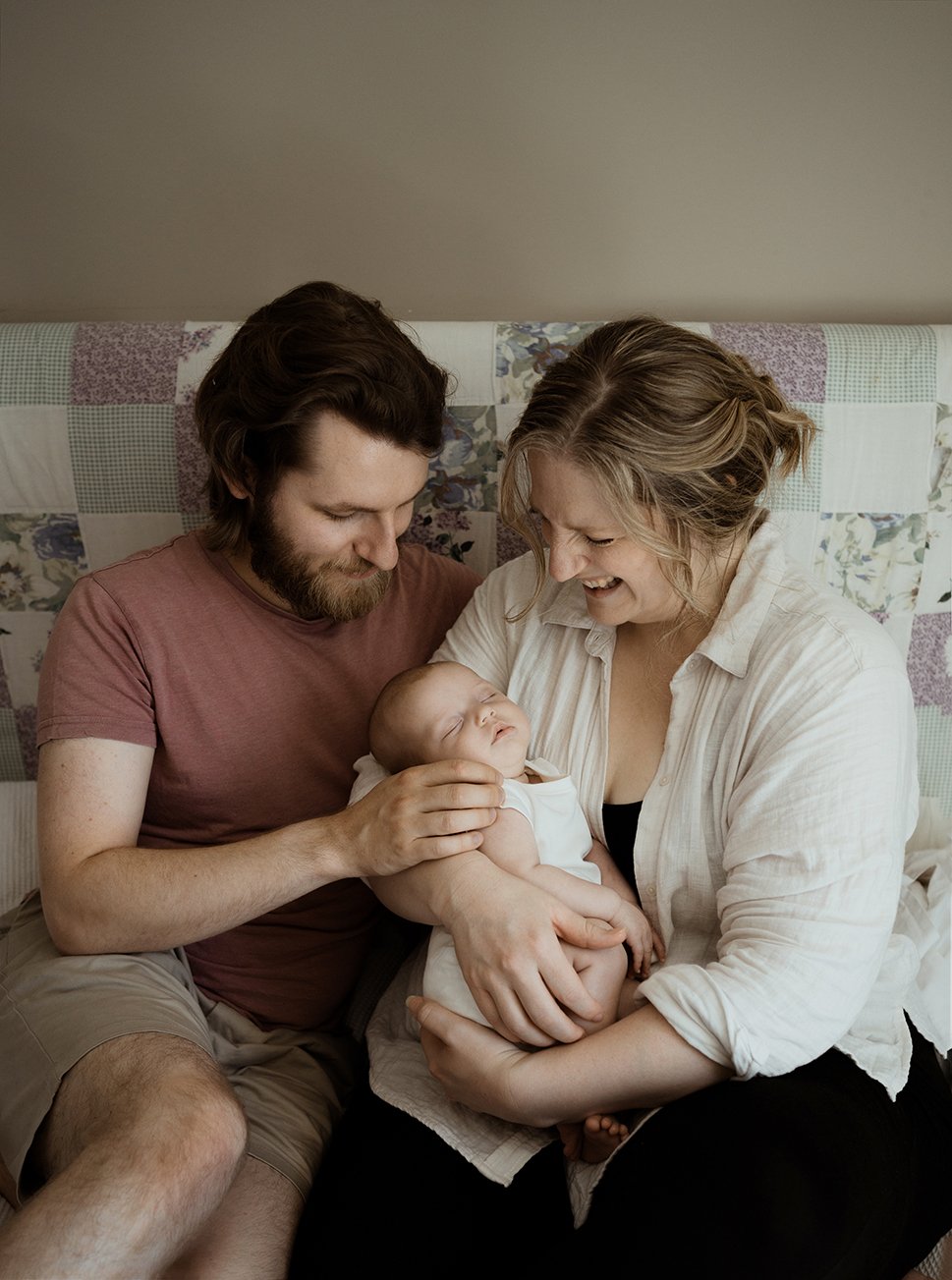 Newborn photographer Bristol - image of a couple with their newborn baby sitting on their bed during a baby photoshoot at their home in Bristol.