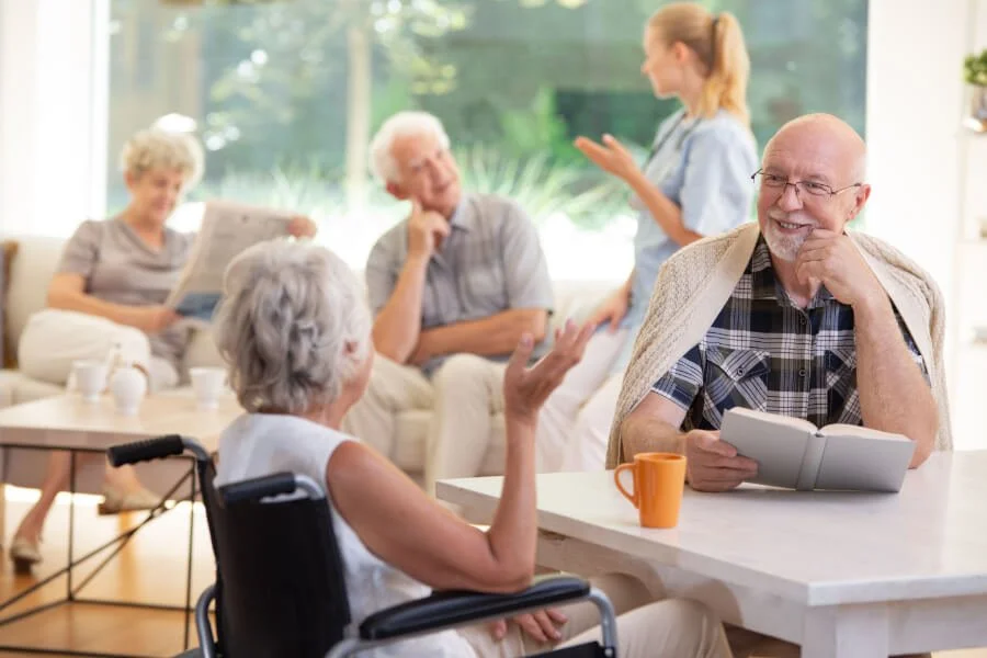 Group of elderly people having a conversation in a bright, sunny living room with large windows. One woman in a wheelchair is talking to a man holding a book, while others are seated in the background, engaged in conversation or reading.