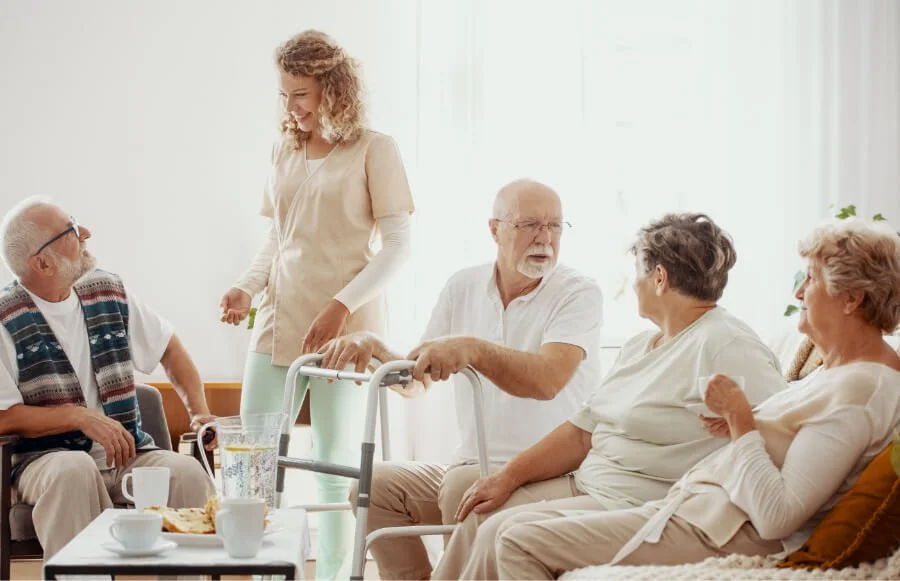 A woman standing and smiling with elderly people sitting on couches and chairs in a bright living room, engaging in conversation.