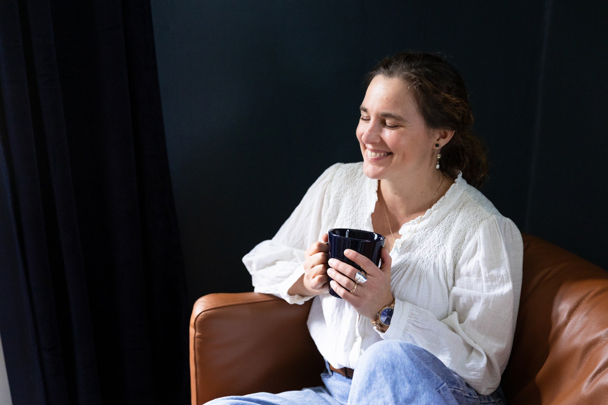 Nainen hymyilee istuen sohvalla, kädessään musta muki.