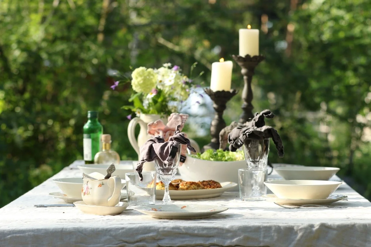 Ulos katettu ruokailupöytä, jossa on kynttilänjalkoja ja kukkakimppu sekä lautasia, juomalaseja ja tarjoiluesineitä, taustalla vihreitä puita.