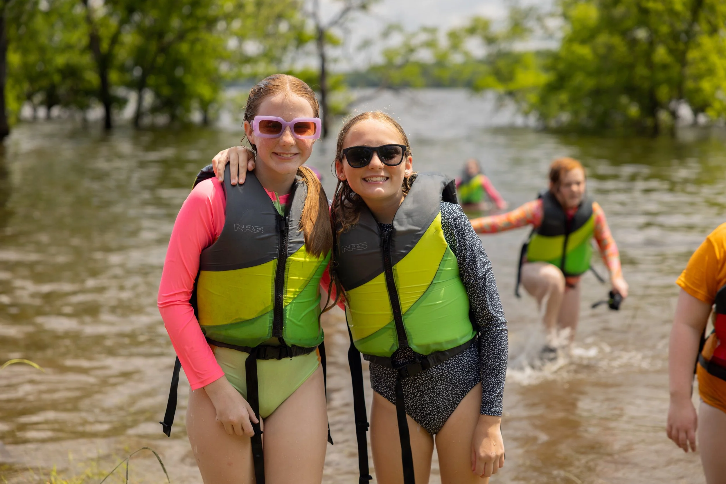 Two young women wearing life jackets and sunglasses standing in the water with others in water behind them, surrounded by green trees.