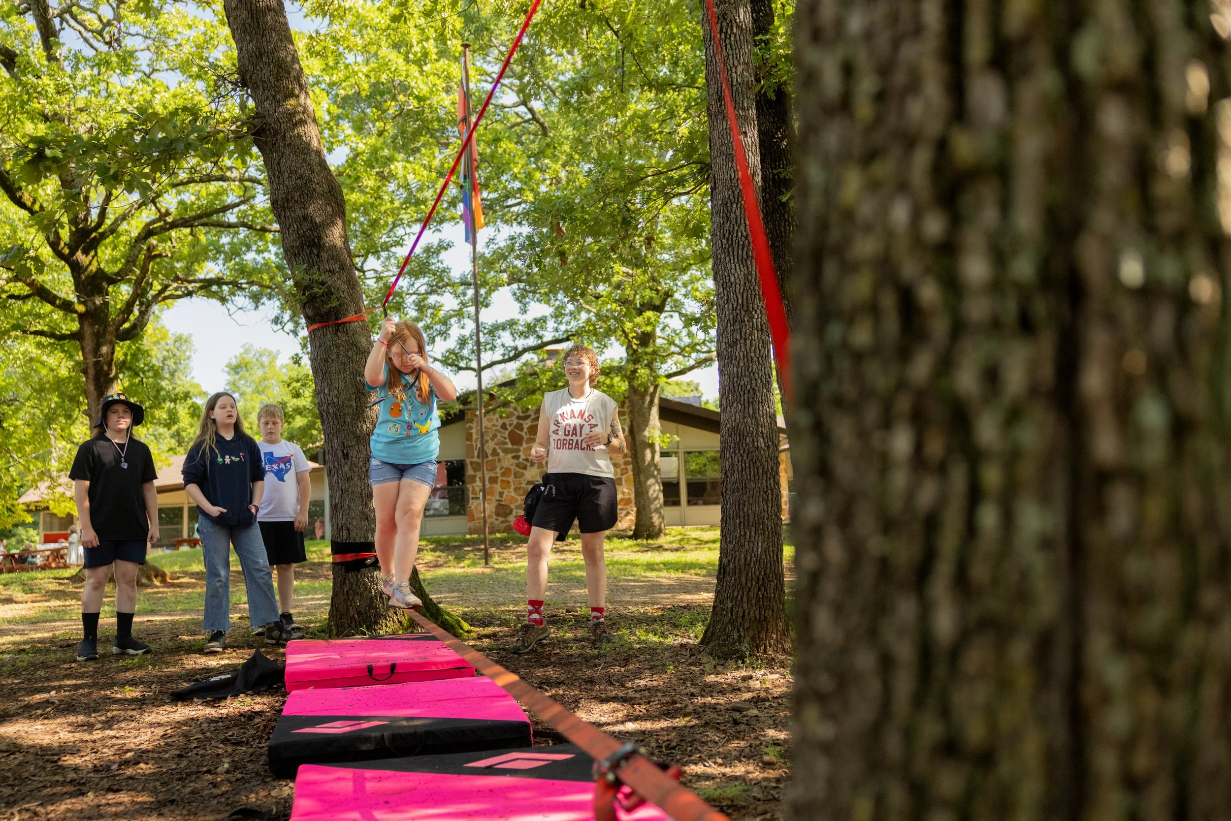 A girl balancing on a tightrope swing between two trees during a camp activity, watched by four children and a counselor in a wooded outdoor area.