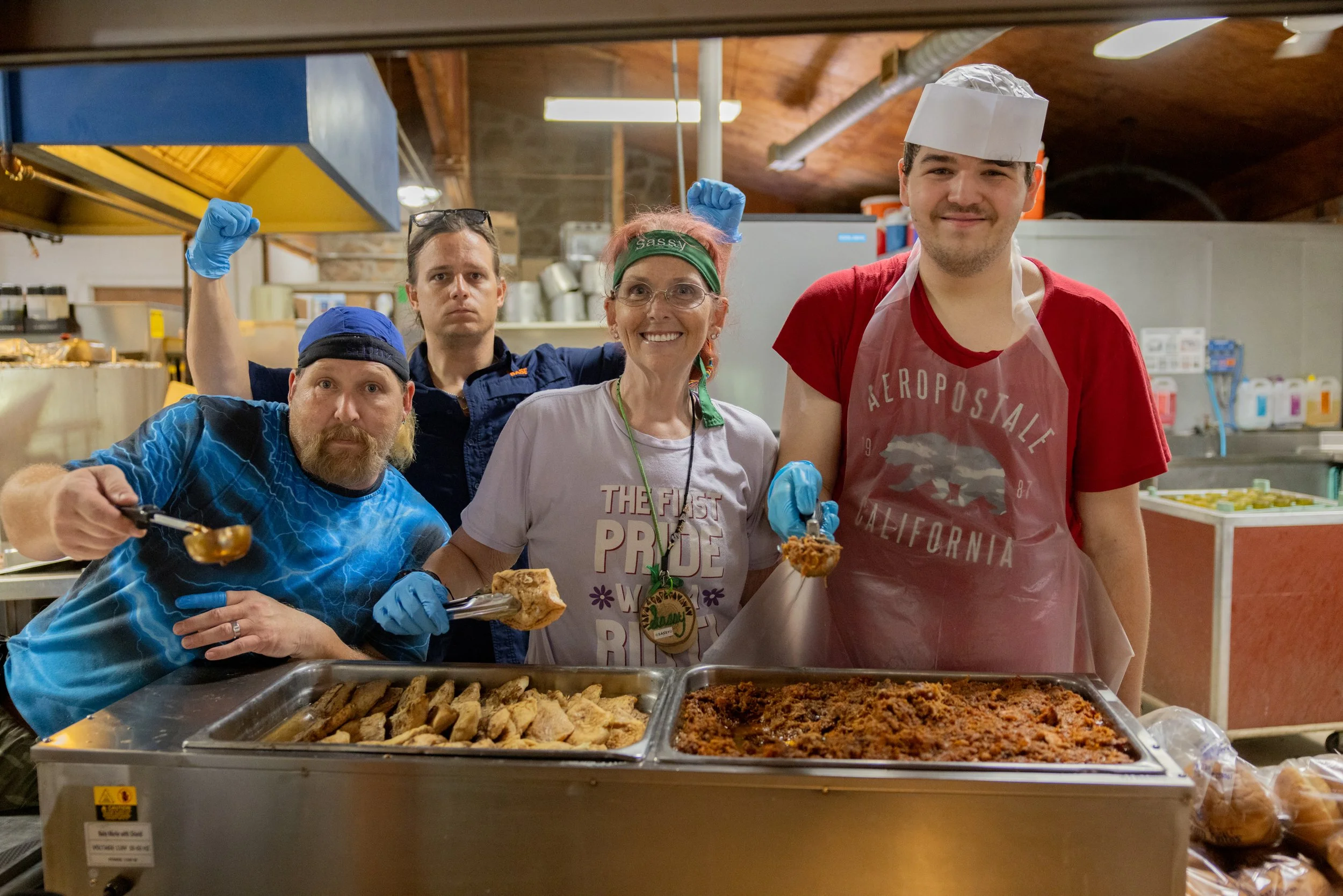 Four people in a kitchen, serving and presenting food, with a tray of baked goods and a pan of baked beans or chili in front of them, smiling and looking at the camera, wearing aprons and gloves.
