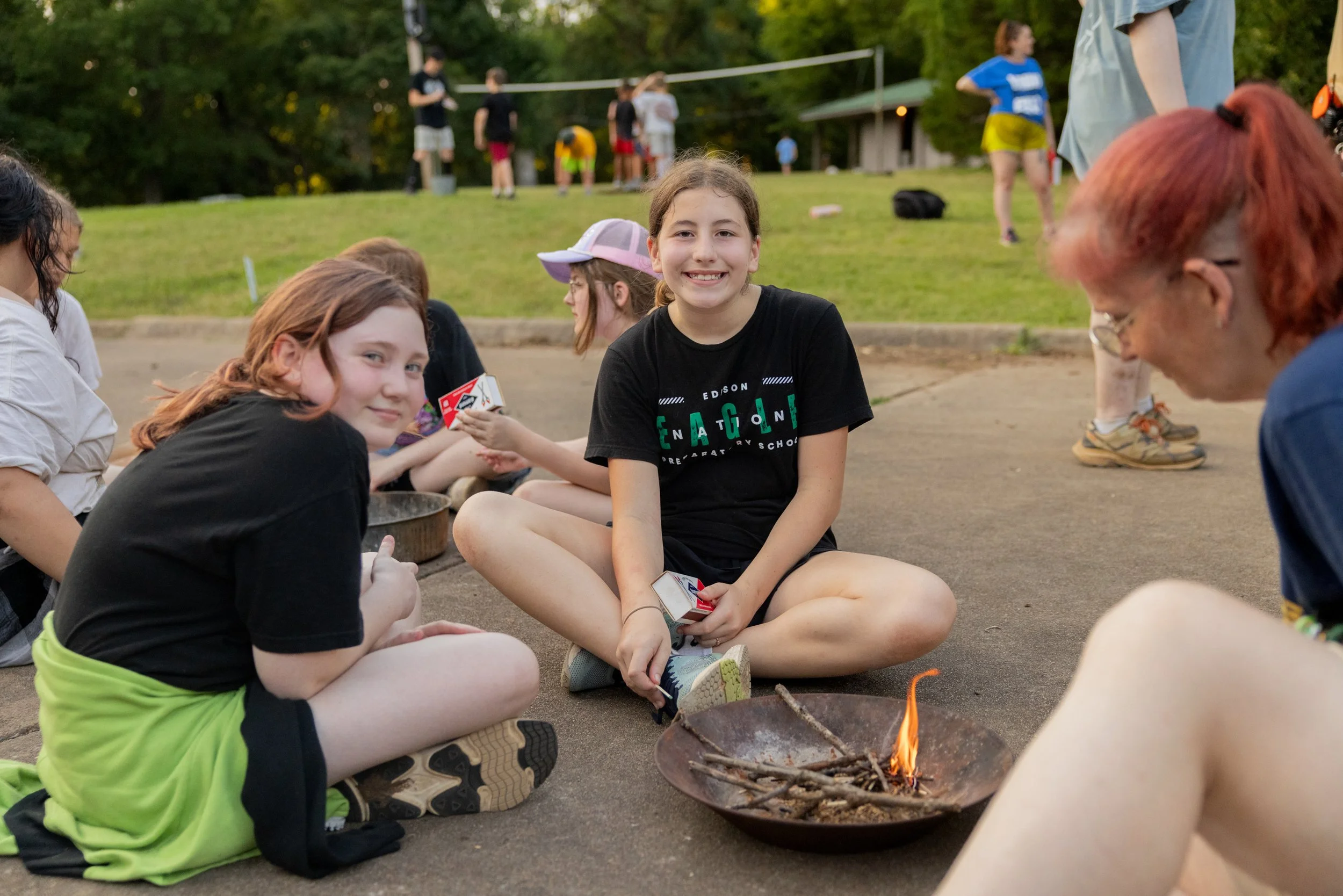 Group of kids sitting around a small campfire outdoors, smiling, with a park or camp setting in the background.
