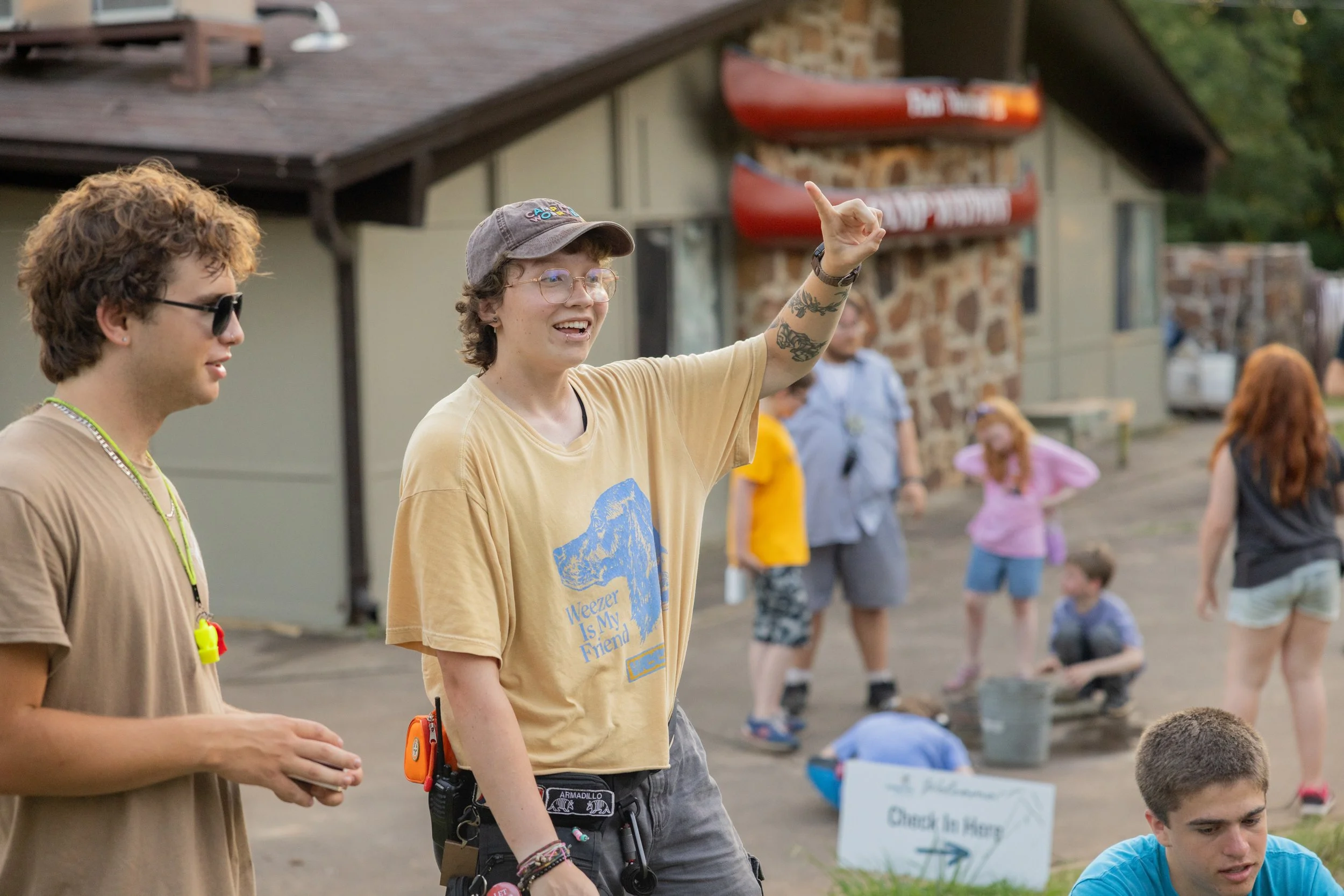 Several people gather outdoors near a building with striped canoes mounted on the wall, some children are involved in activities while adults supervise.