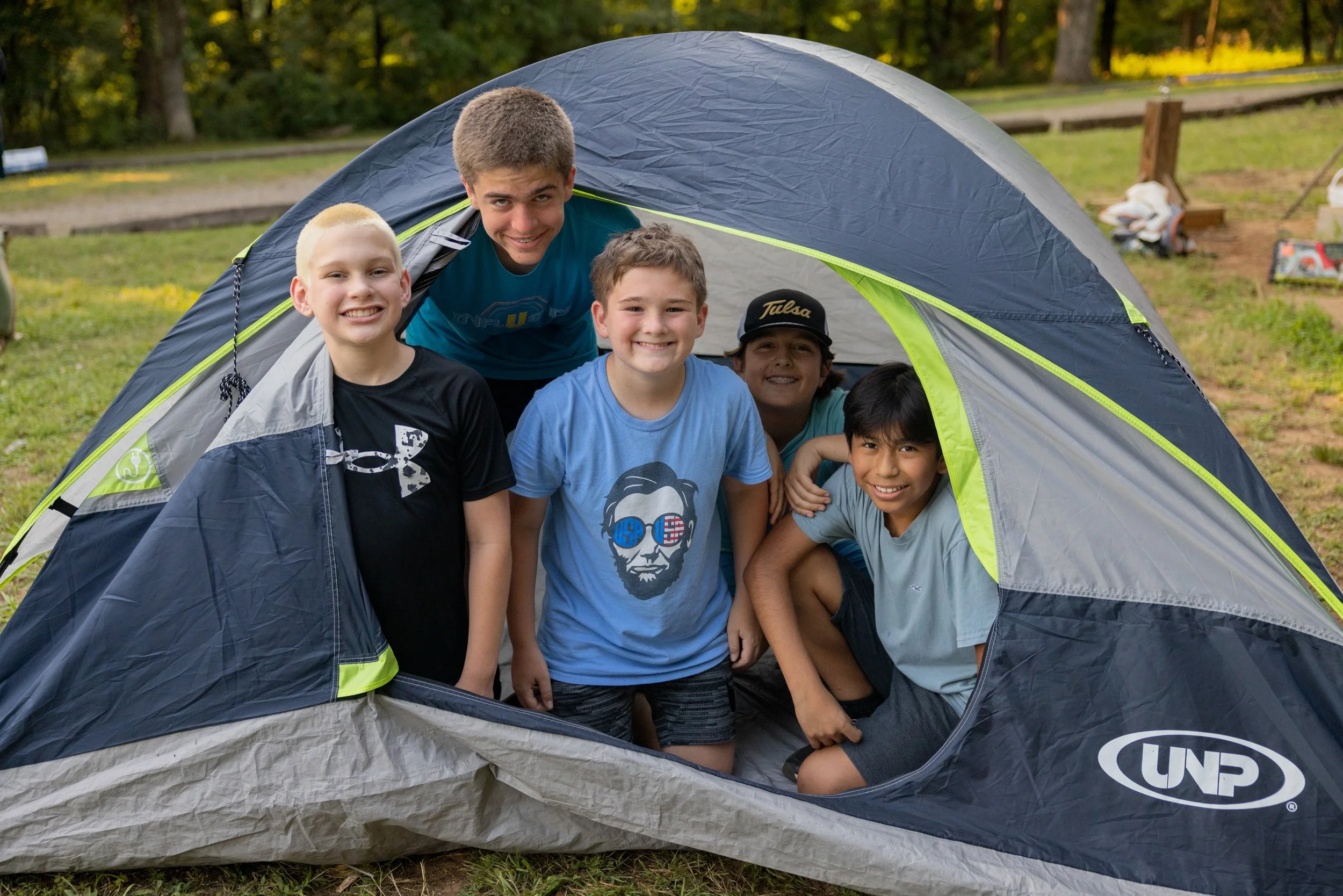 Six boys are smiling and posing inside an open tent outdoors with a grassy area and trees in the background.