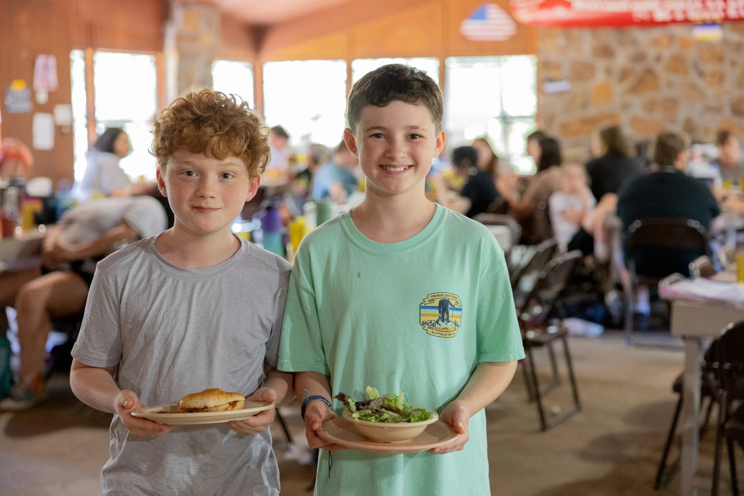 Two young boys standing indoors at a gathering, each holding a plate of food, smiling at the camera, with a busy room filled with people at tables in the background.