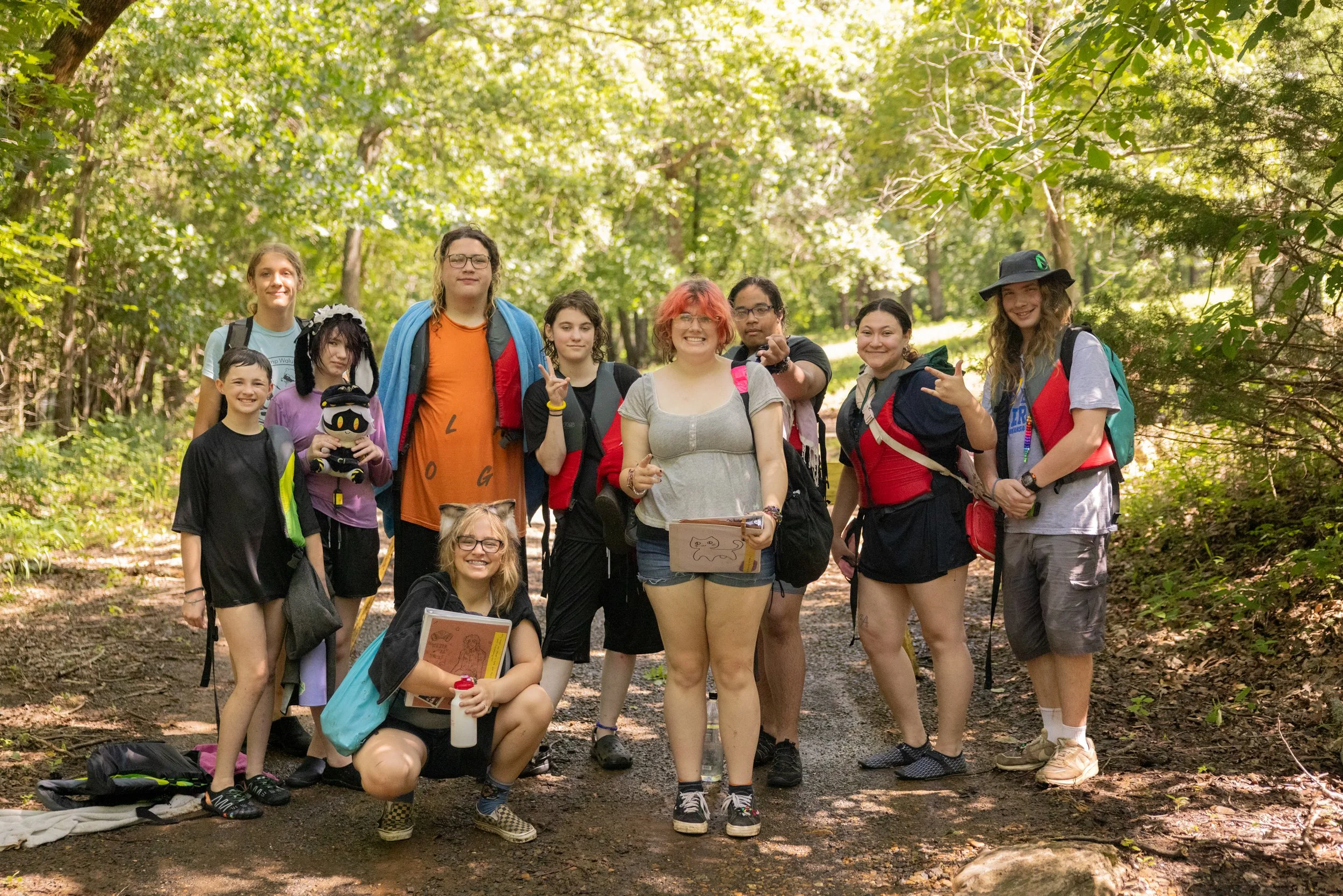 Group of ten people, mostly young women, standing on a forest trail surrounded by trees and greenery, smiling and posing for a photo during daytime.