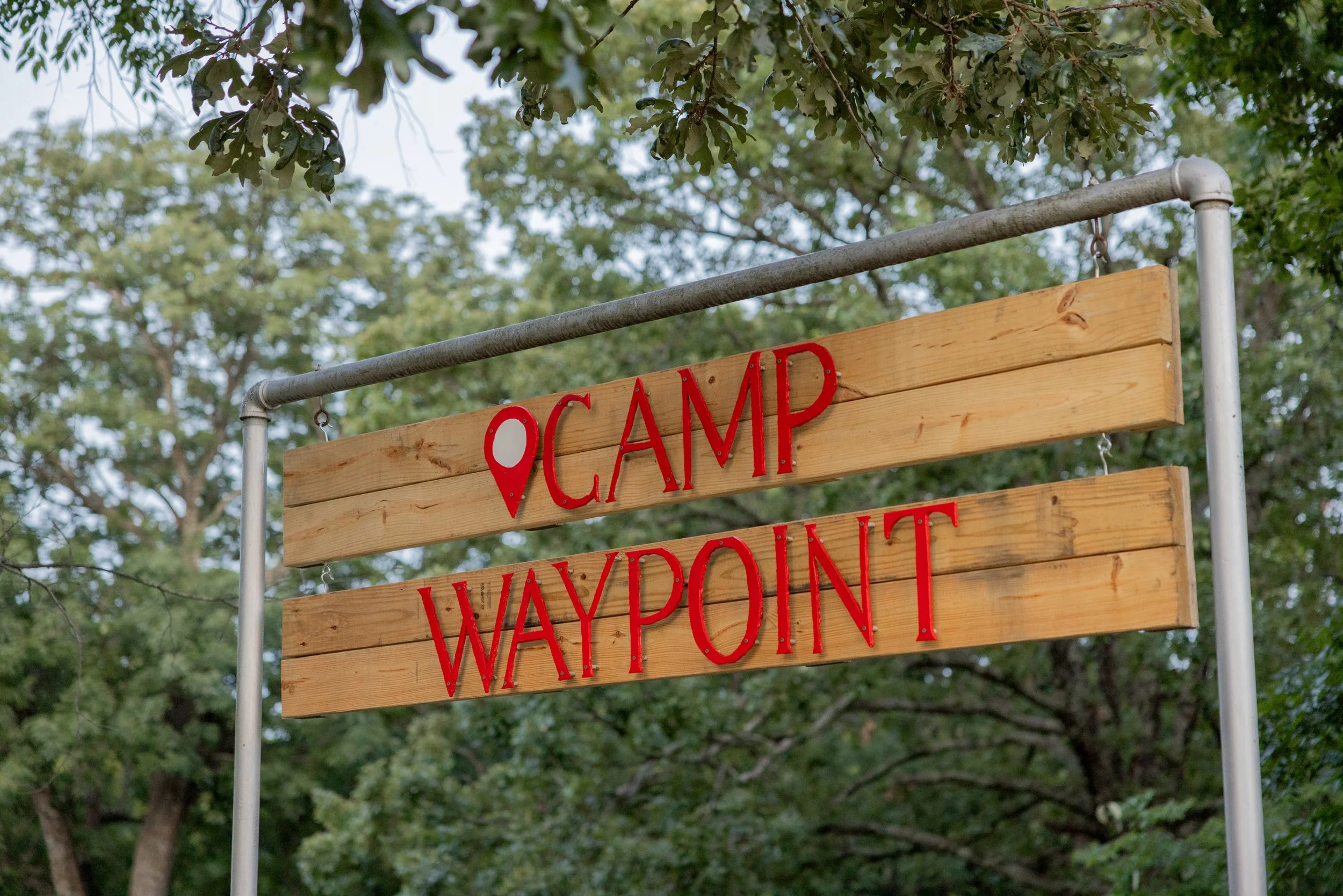Wooden sign with red lettering that reads 'CAMP WAYPOINT' and a red location pin icon, hanging from a metal frame, with trees and sky in the background.