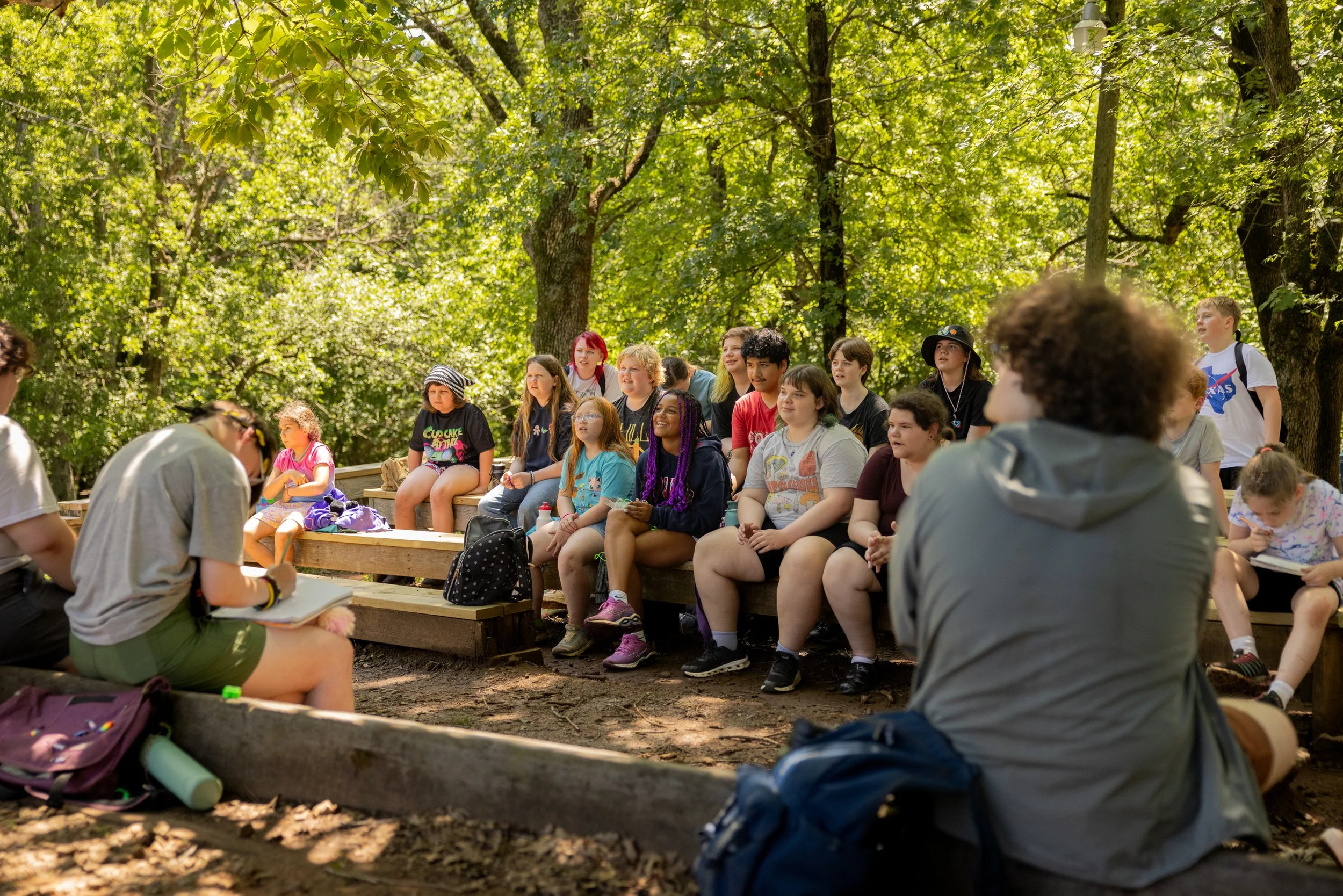 Group of children and young adults sitting on wooden benches outdoors in a lush, green forest, listening to a speaker or teacher.