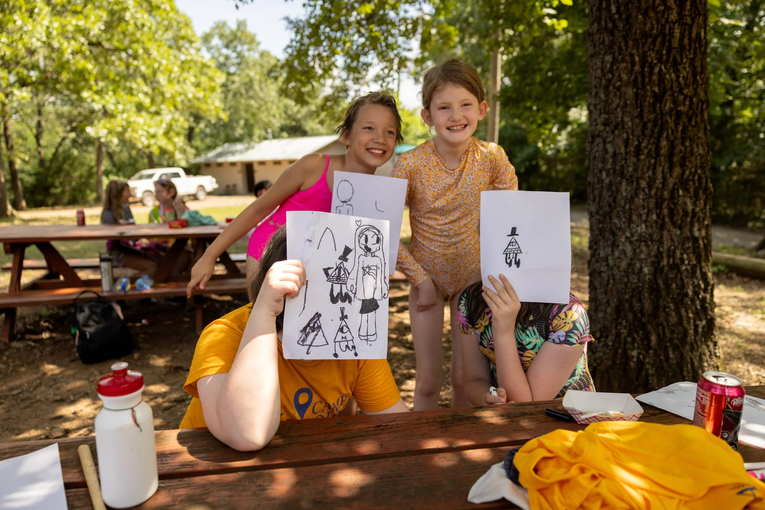 Three children smiling at a picnic table outdoors, one holding up a drawing, two covering their faces with papers, with others in the background at a picnic area under trees.