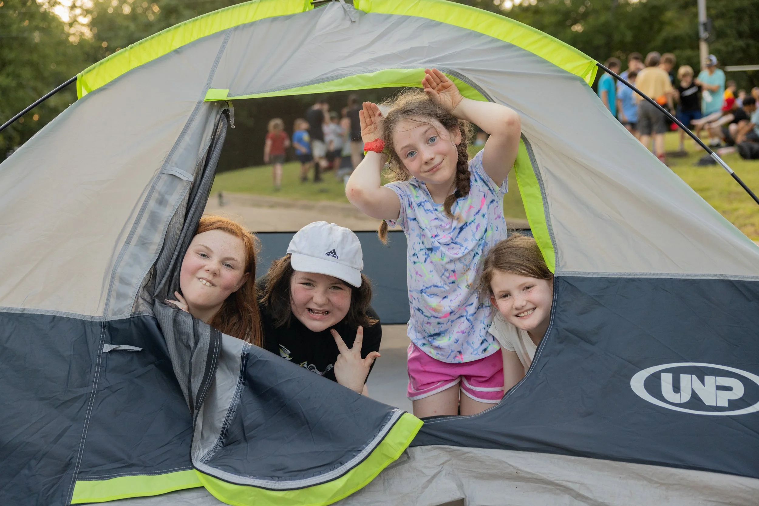 Four girls are peeking out and playing inside a tent outdoors at a campsite with other people in the background.