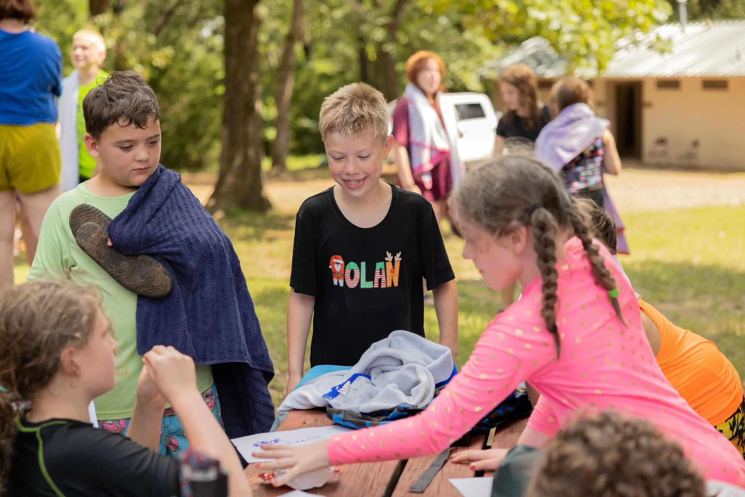 Children gathered around a picnic table outdoors, with some standing and some sitting, engaged in an activity, surrounded by trees and a building in the background.