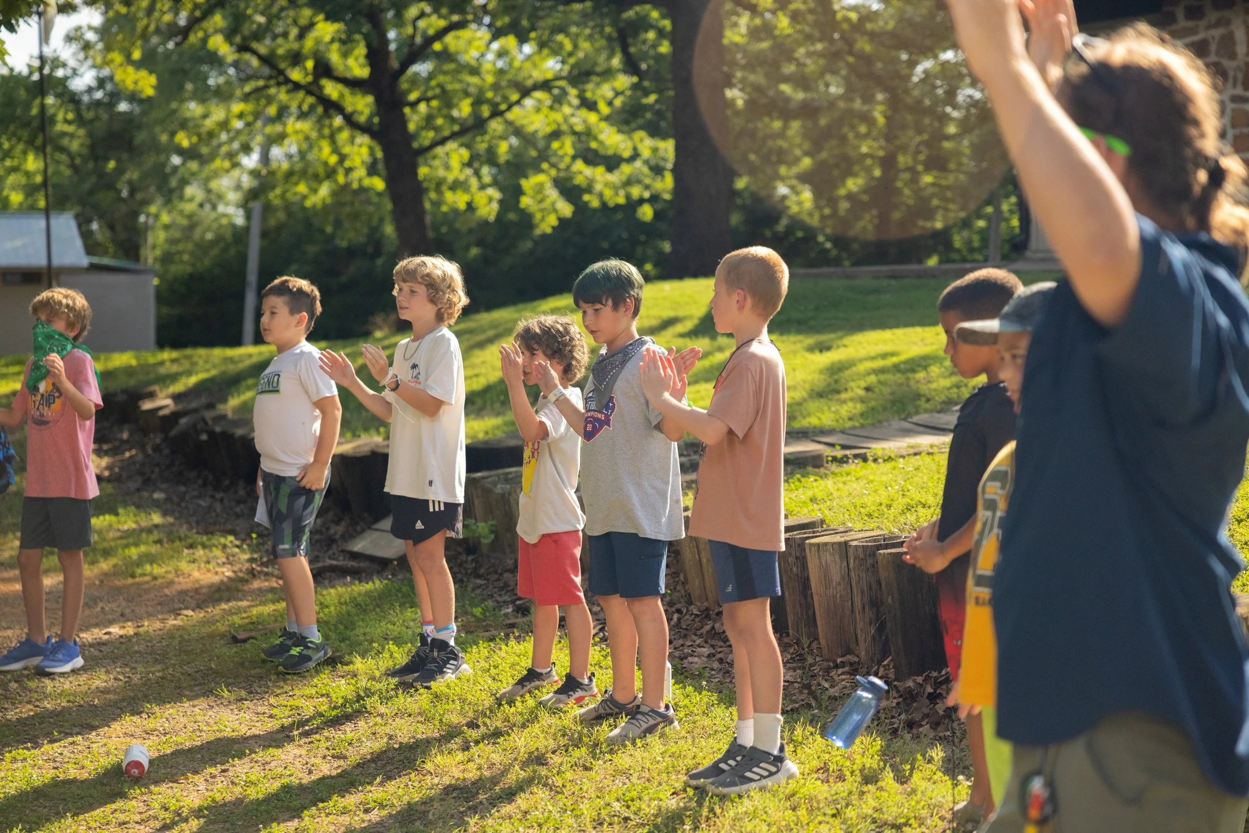 Children standing outdoors in a line, clapping, with a teacher or adult supervising, on a grassy area with trees in the background.