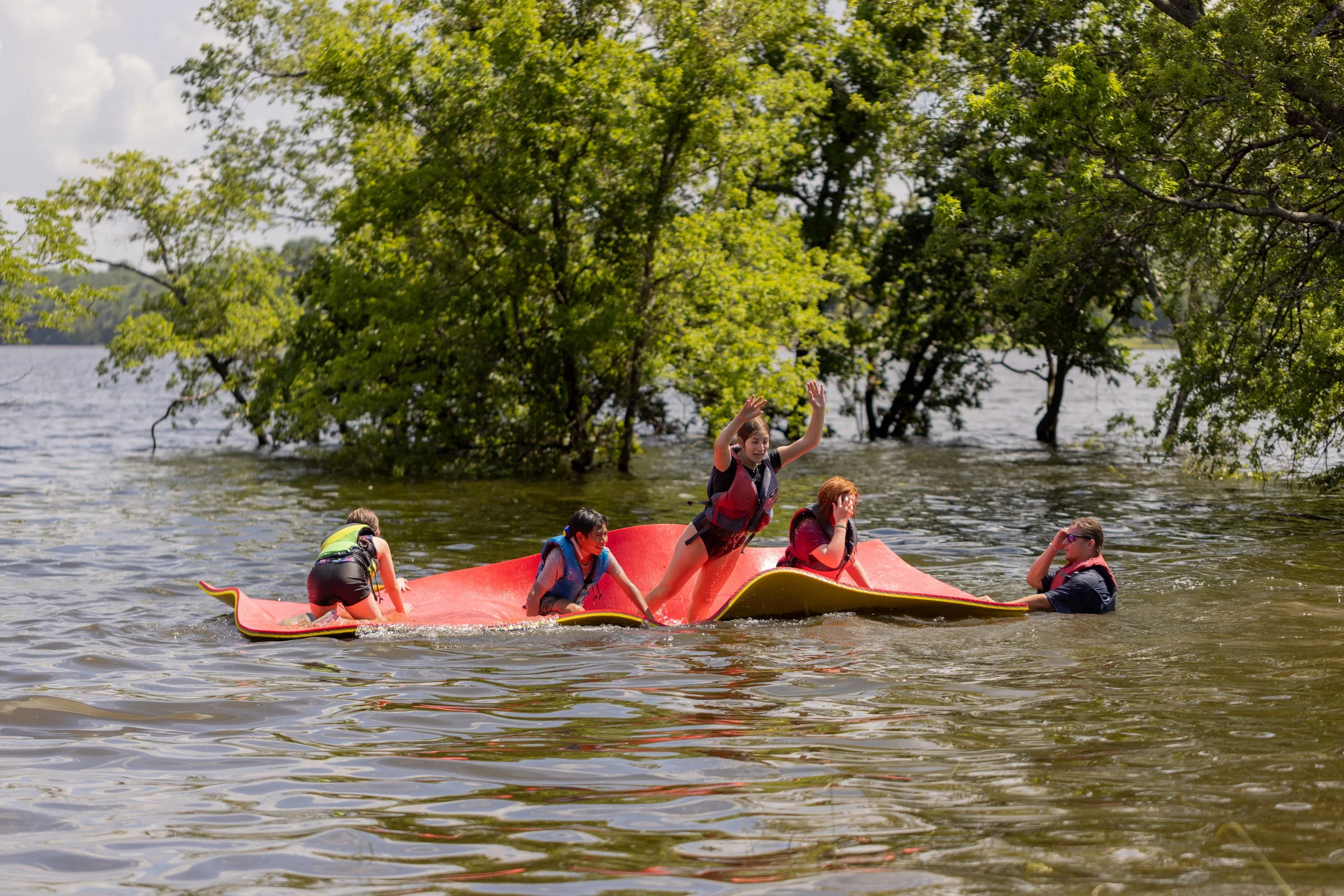 Group of children on a partially submerged blue and red inflatable raft in a lake, surrounded by green trees, with one child standing and raising arms, and others sitting or lying, wearing life jackets.