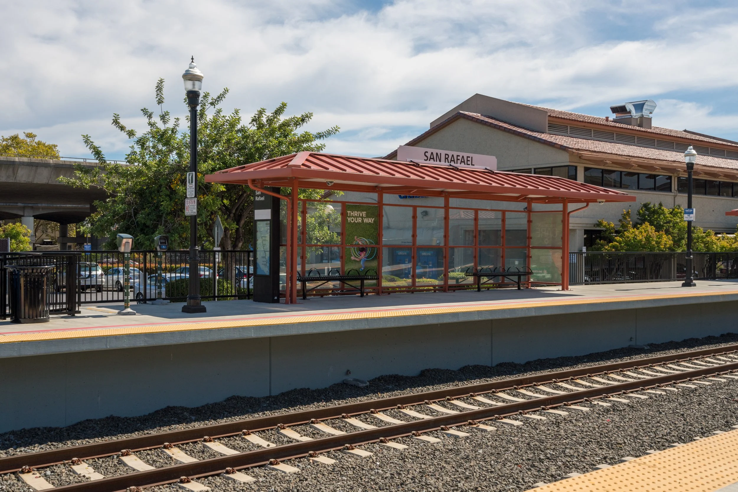 Empty train station platform with a red shelter, benches, lampposts, parking lot, and a building with a sign reading 'San Rafael' in the background.