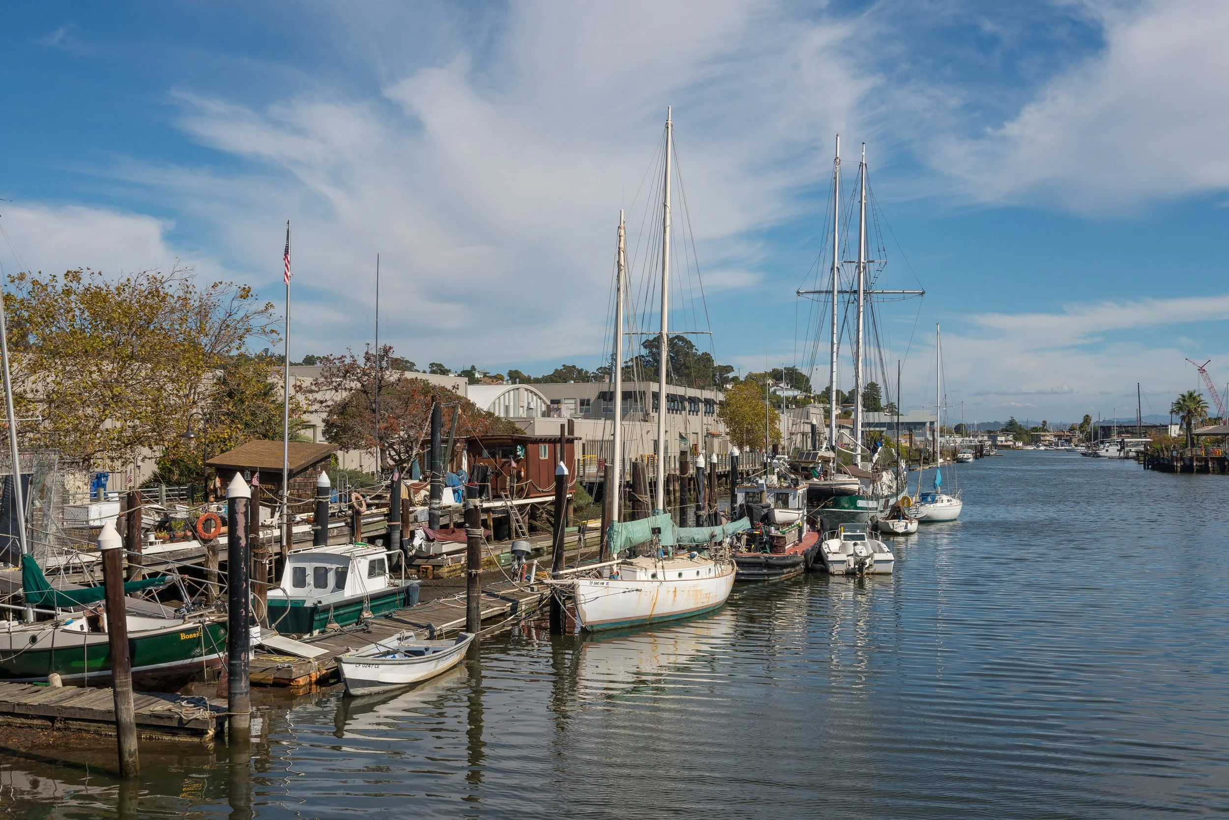 A marina with boats docked along wooden piers, buildings and trees in the background, under a partly cloudy sky.