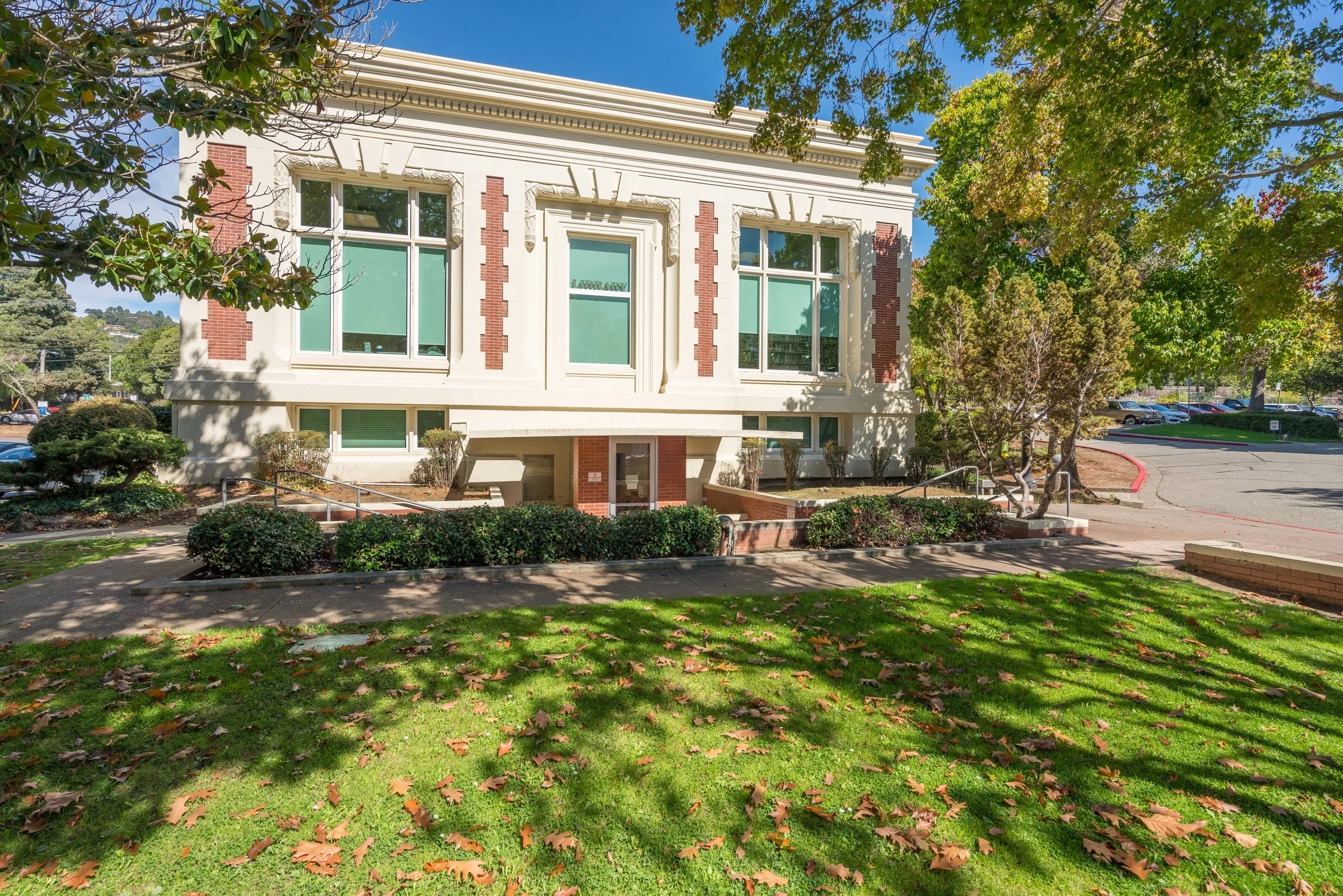 A two-story building with cream and red brick exterior, three large windows on the upper floor, surrounded by green trees and a grassy lawn with fallen leaves.