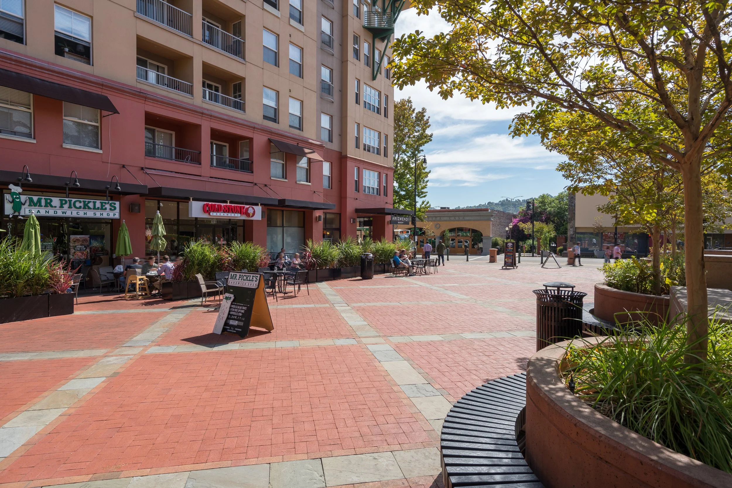 An outdoor plaza with a pizzeria called Mr. Pickle's and a Cold Stone ice cream shop, featuring tables, umbrellas, and people walking and sitting.