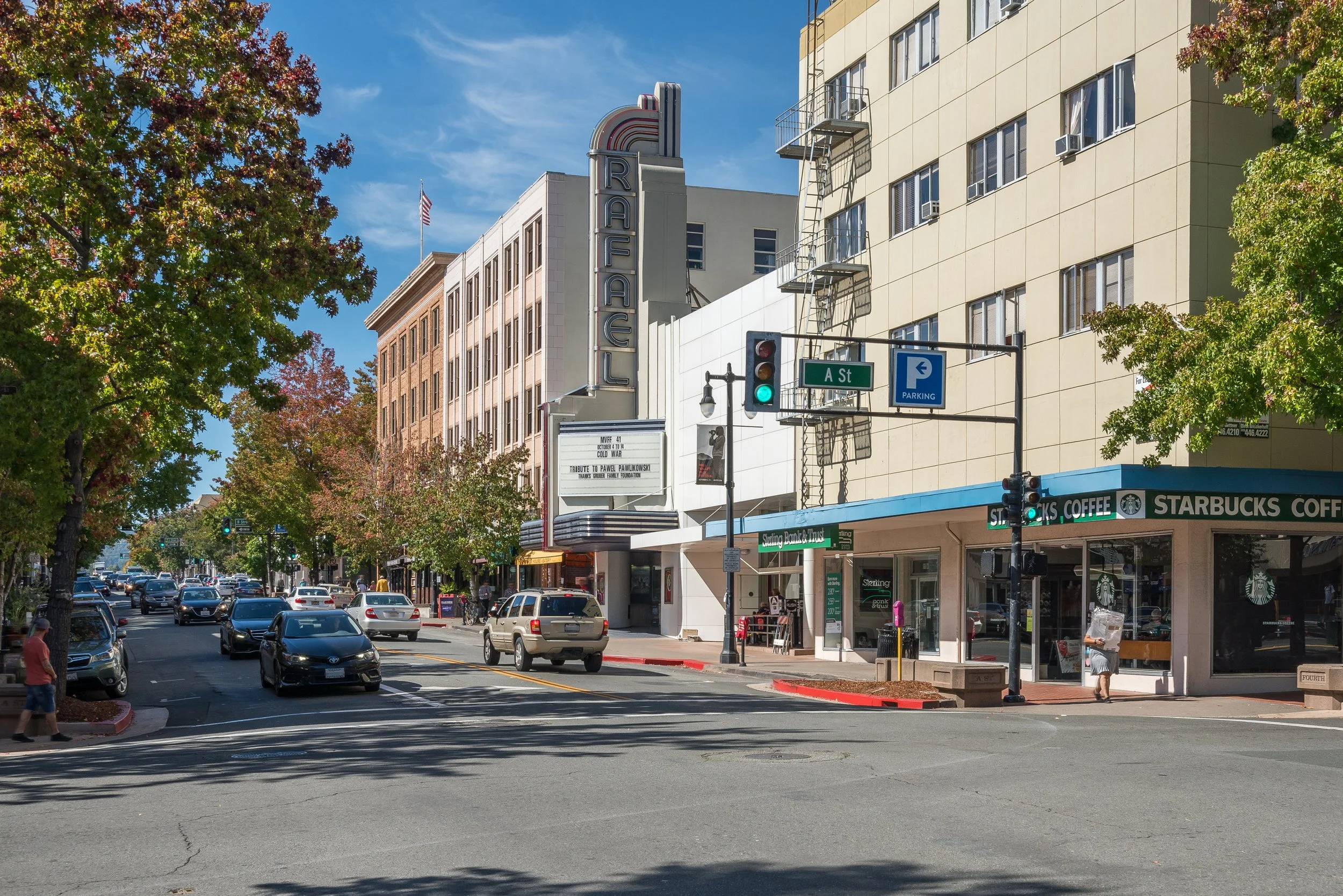 City street corner with old cinema building, Starbucks coffee shop, parked cars, trees, and traffic lights, under a clear blue sky.
