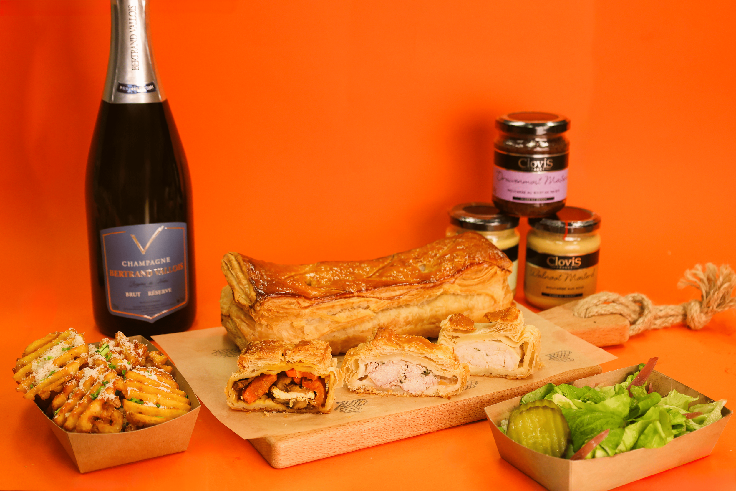 A variety of food items including a bottle of champagne, a puff pastry dish with chicken, a side of waffle fries, jars of jam, and a fresh green salad on an orange background.