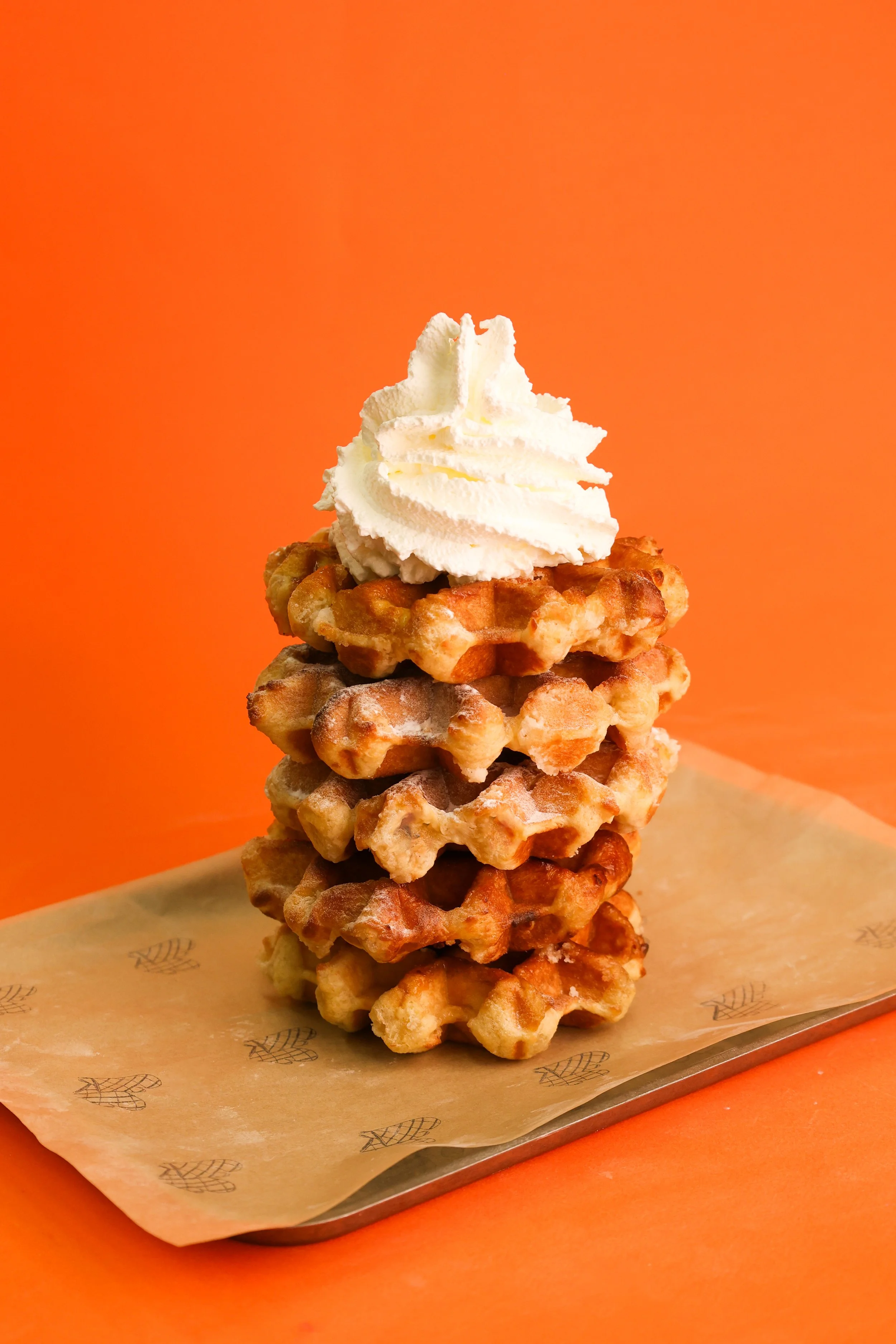 A stack of five waffles topped with whipped cream on an orange background, placed on a paper-lined tray.